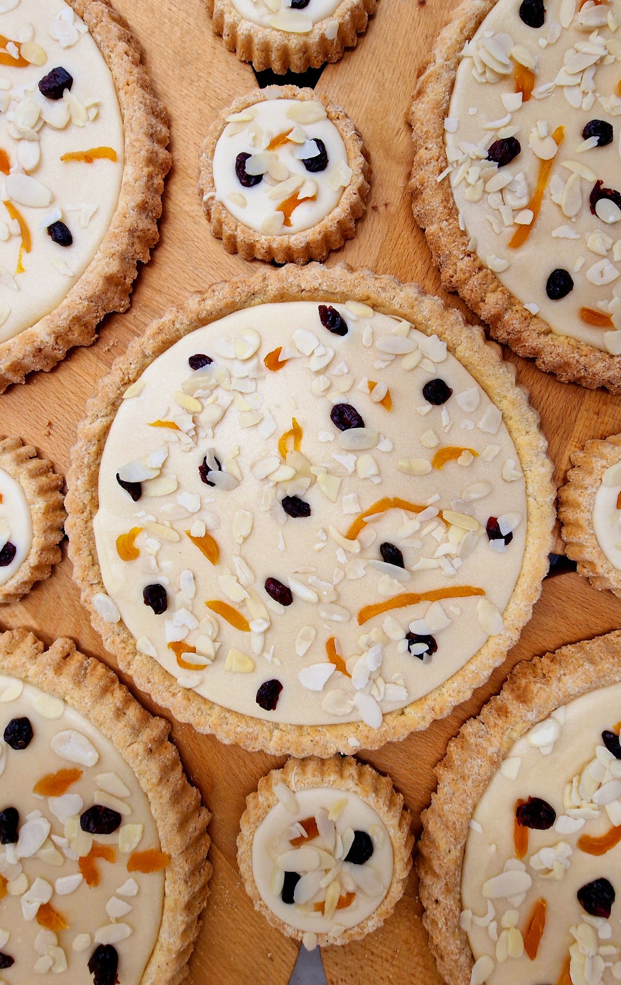 Tarty z kremem, ozdobione migdałami, suszonymi owocami i rodzynkami, ułożone na drewnianym tle.