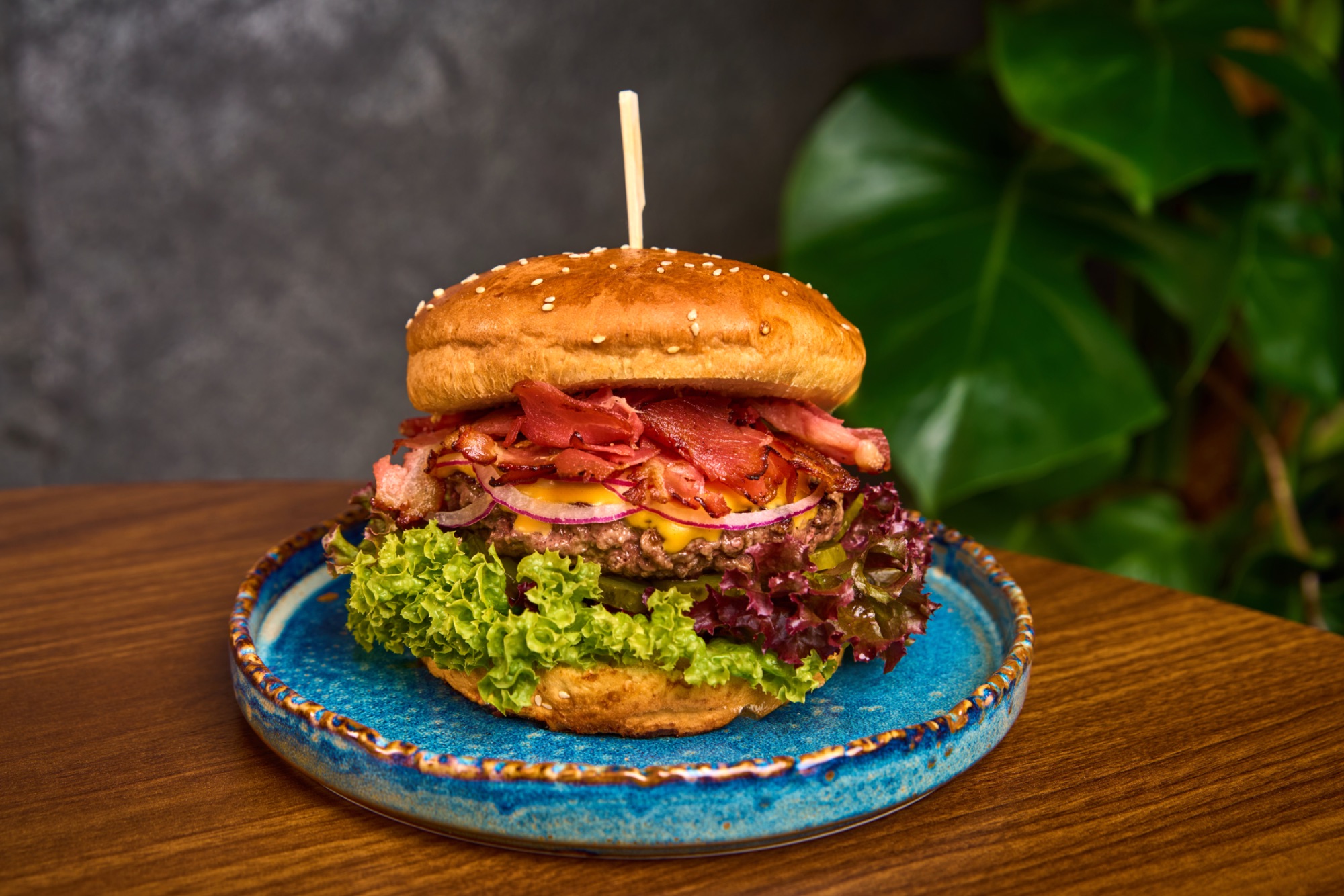 Burger z soczystym mięsem, serem cheddar, sałatą i wędzoną szynką na talerzu, w tle liście roślin.