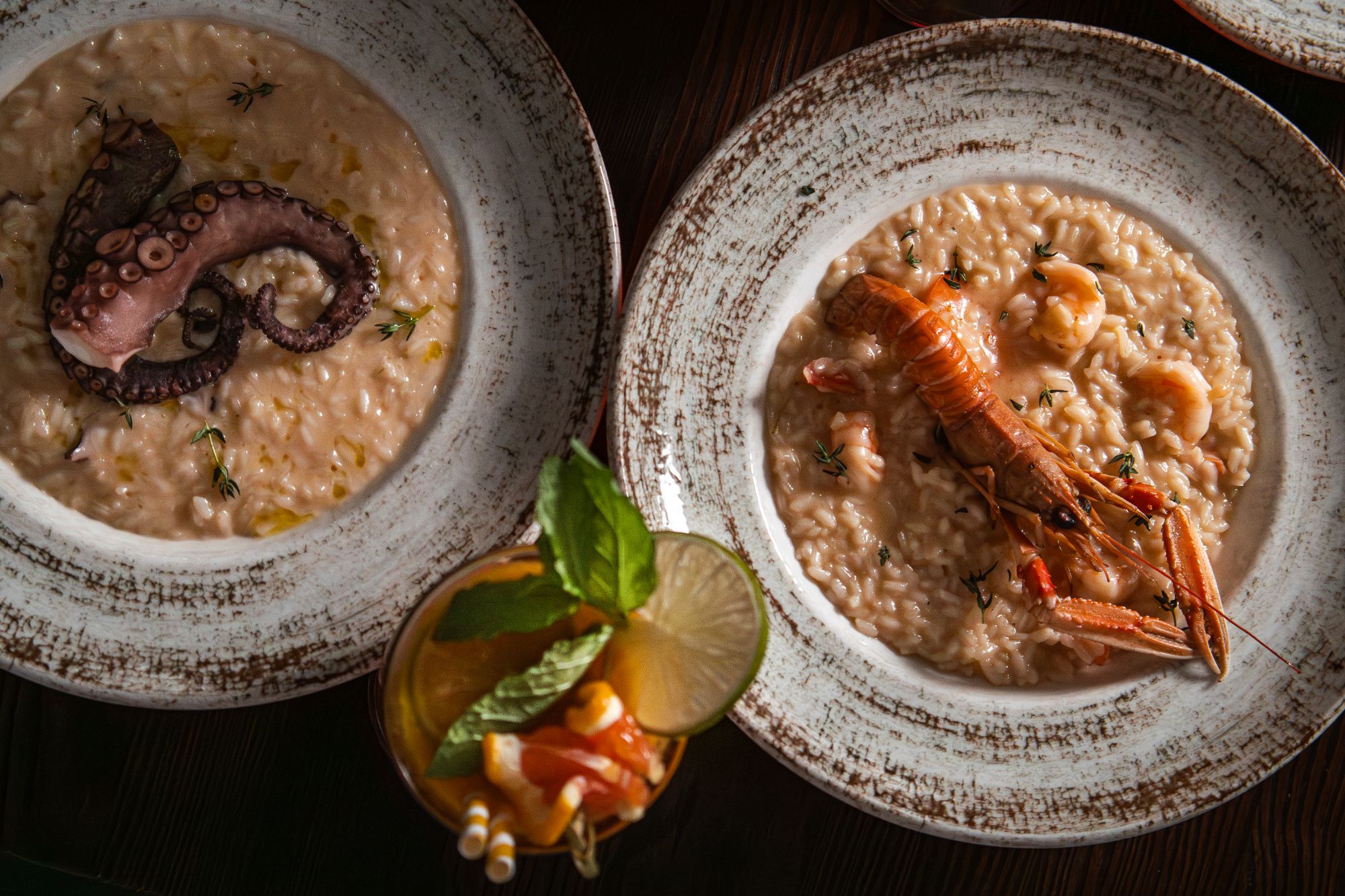 Risotto z krewetkami i ośmiornicą podane w białych talerzach, obok kolorowy koktajl owocowy.