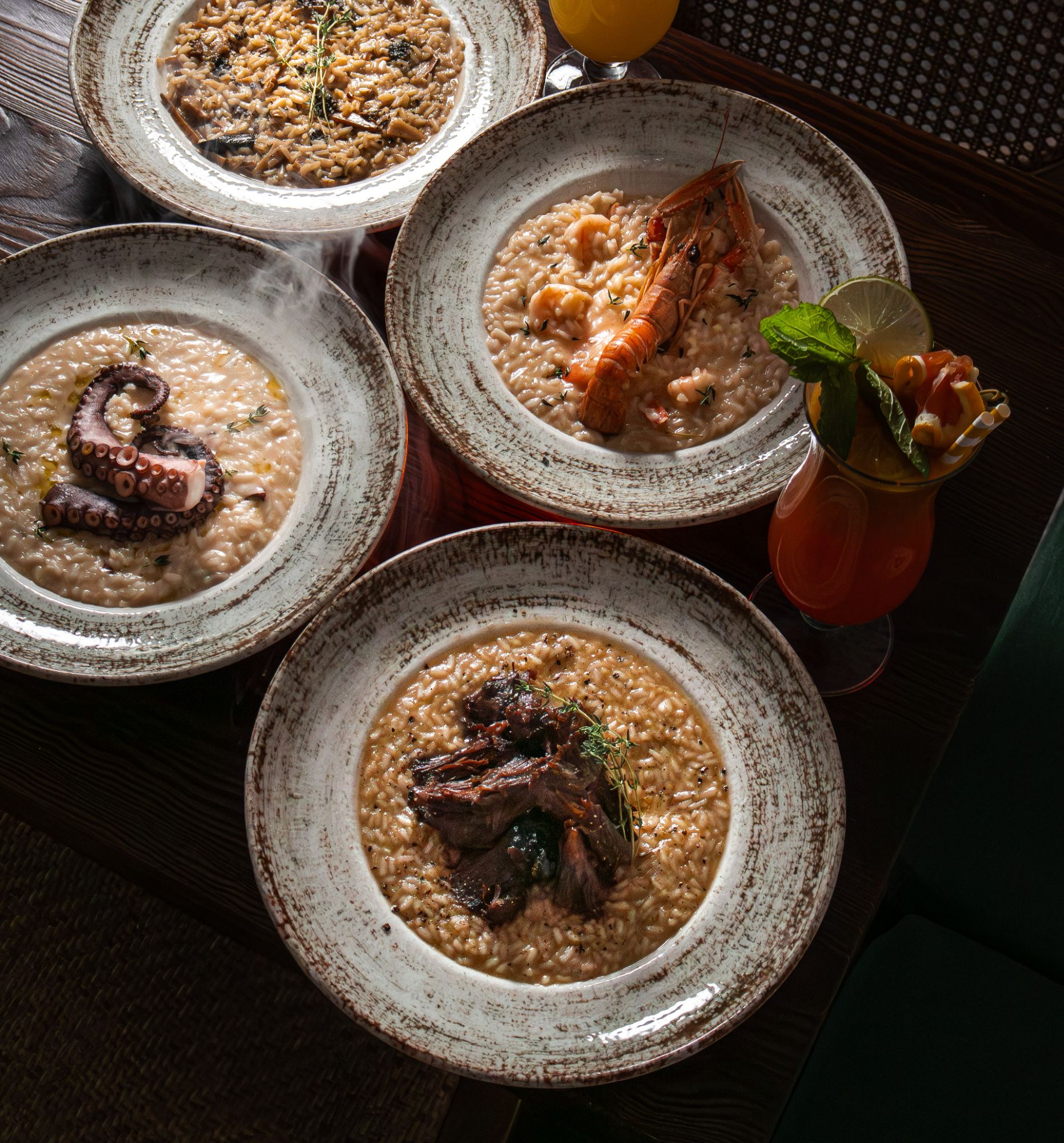 Risotto z owocami morza, krewetkami, ośmiornicą i mięsem, podane w eleganckich talerzach, obok koktajl.