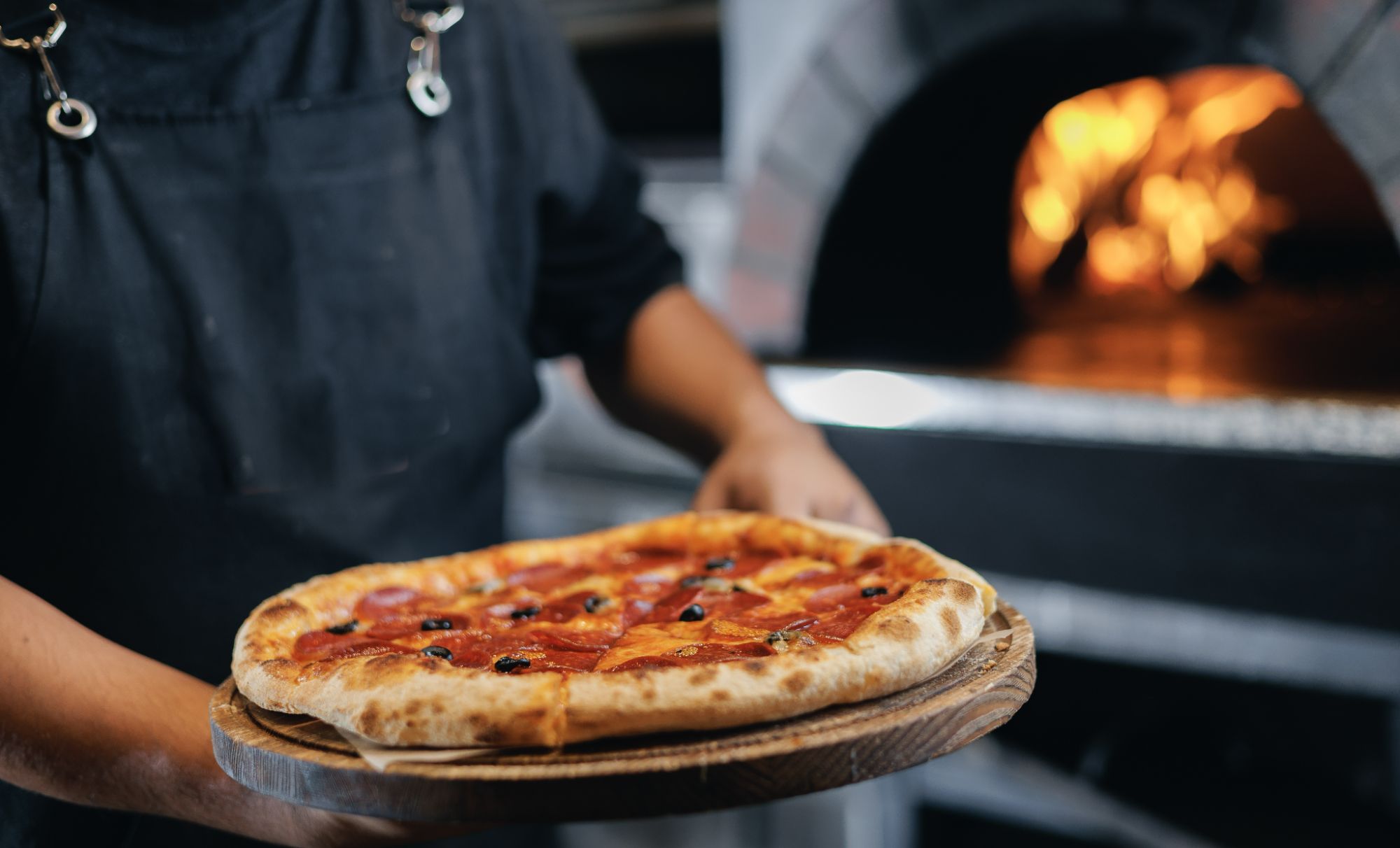 Osoba podaje świeżo upieczoną pizzę na drewnianym talerzu, w tle widać piec opalany drewnem.