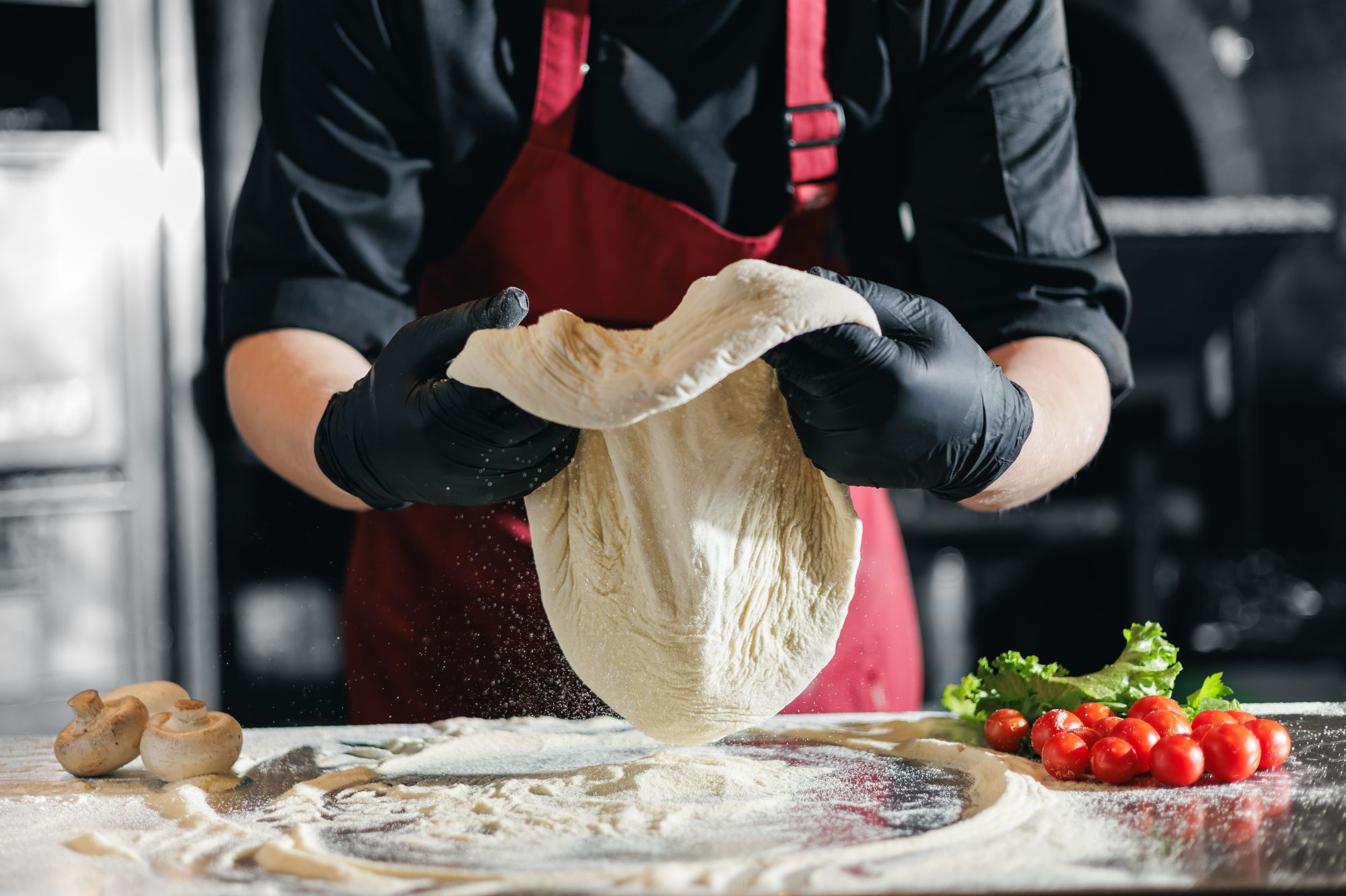 Kucharz w rękawiczkach wyrabia ciasto na pizzę na blacie kuchennym z pomidorami i sałatą.