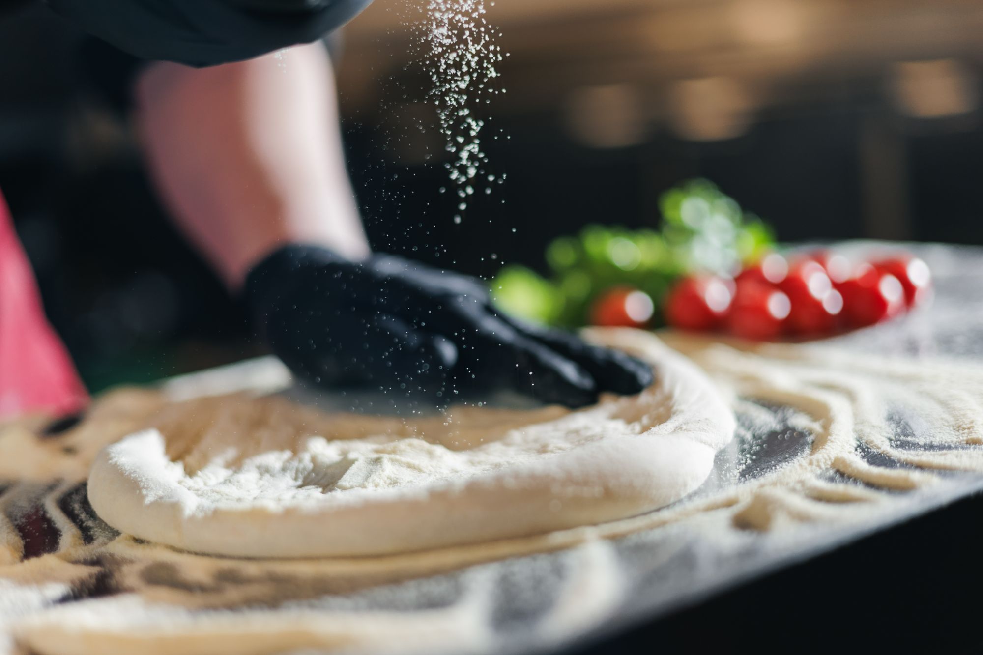 Osoba w czarnych rękawiczkach posypuje mąkę na cieście na pizzę obok pomidorów i świeżej bazylii.