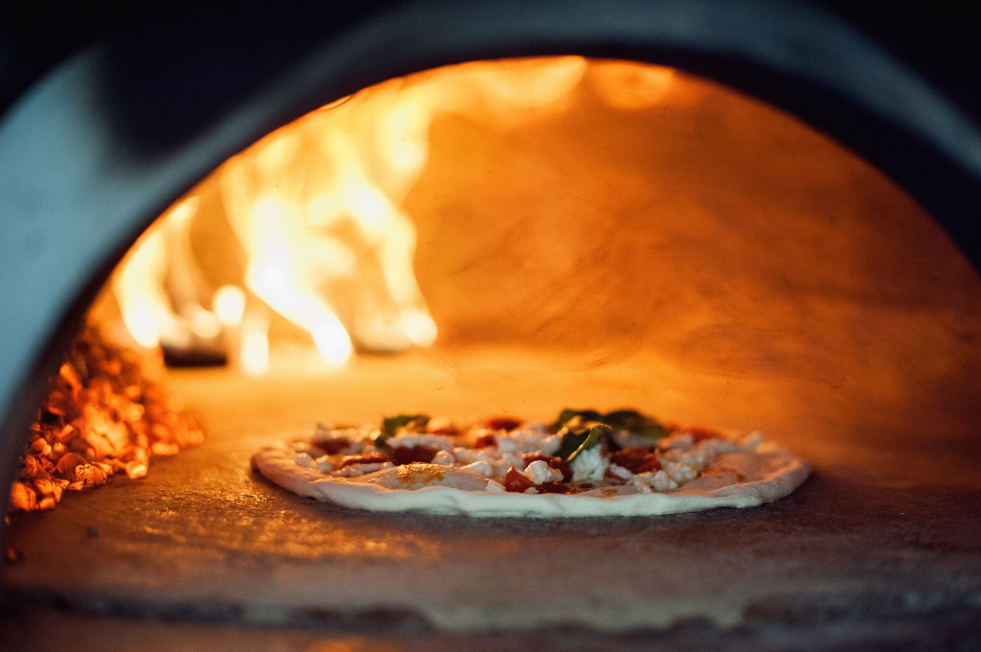 Pizza pie baking in a wood-fired oven, with flames and embers visible in the background.