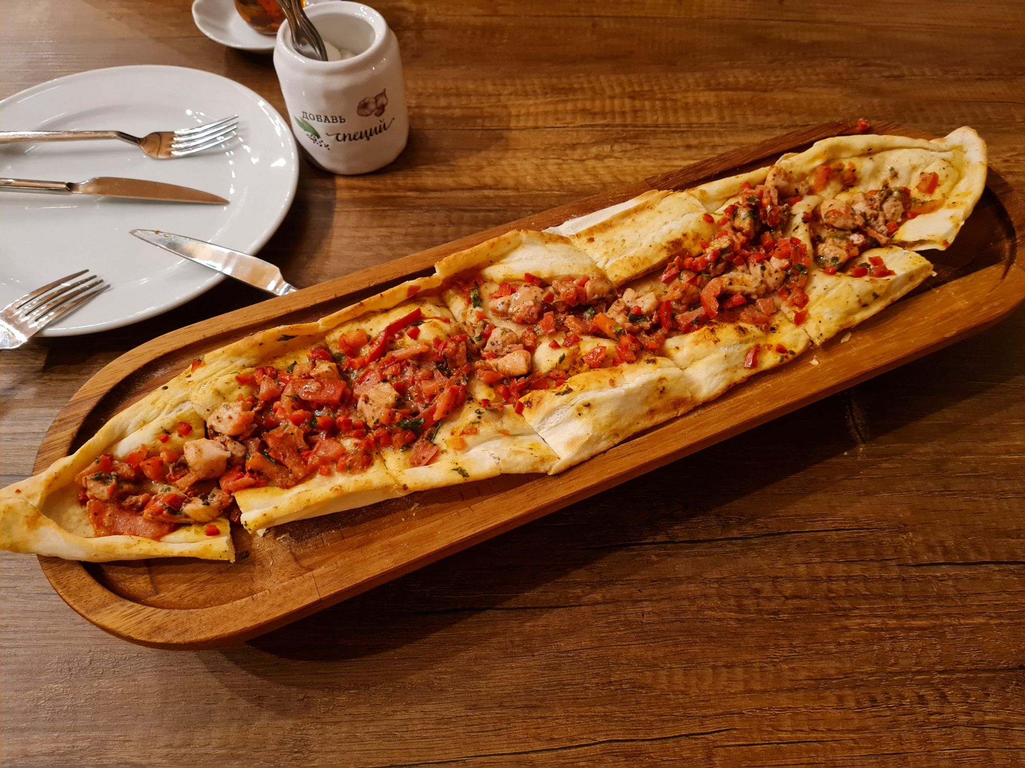 Długa pizza na drewnianej desce, pokryta mięsem, pomidorami i przyprawami, w restauracyjnym wnętrzu.