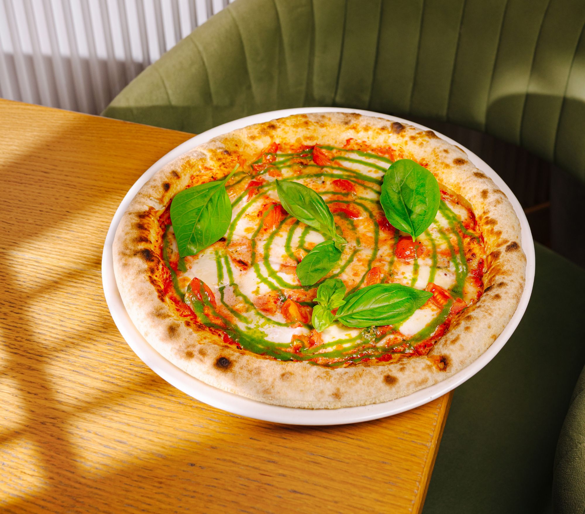 Pizza z sosem pesto, pomidorami i świeżą bazylią na białym cieście, podana na drewnianym stole.