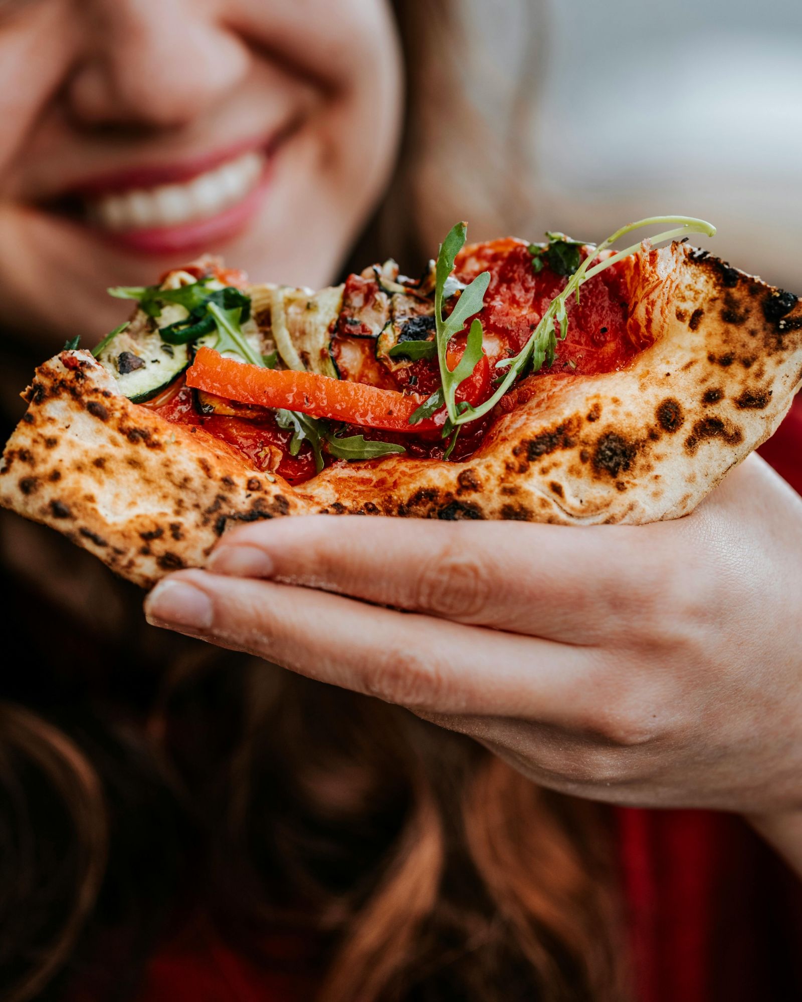Kobieta trzymająca kawałek pizzy z warzywami i ziołami, uśmiechając się w tle.