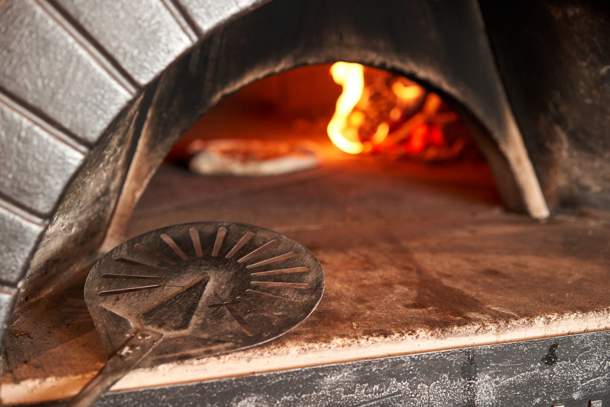 Wnętrze pieca do pizzy z łopatką do pizzy i płonącym ogniem w tle.
