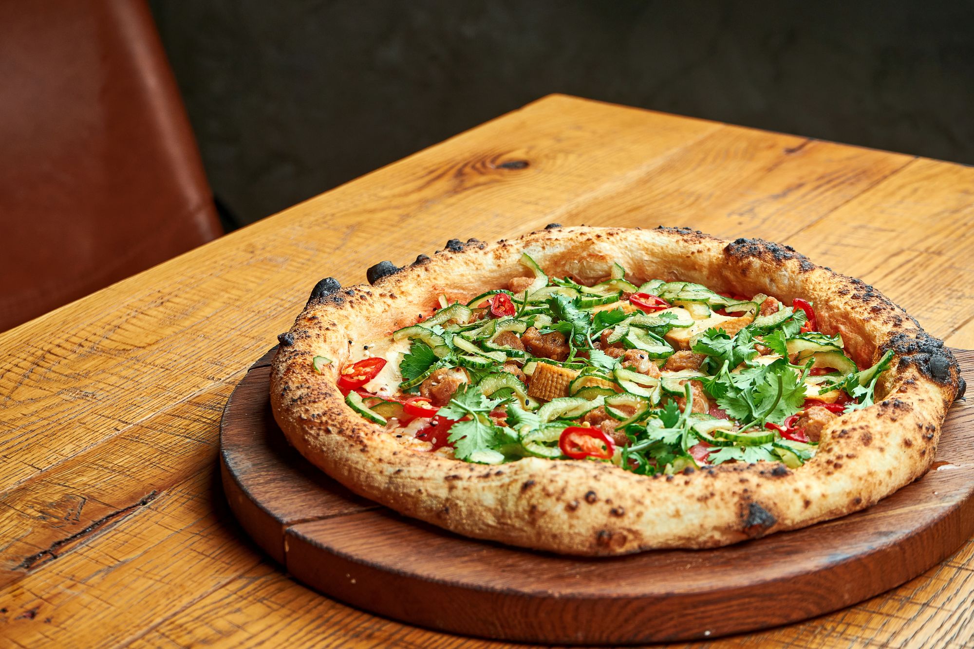 Pizza z rukolą, pomidorami i ziołami na drewnianej desce, leży na stole z naturalnego drewna.