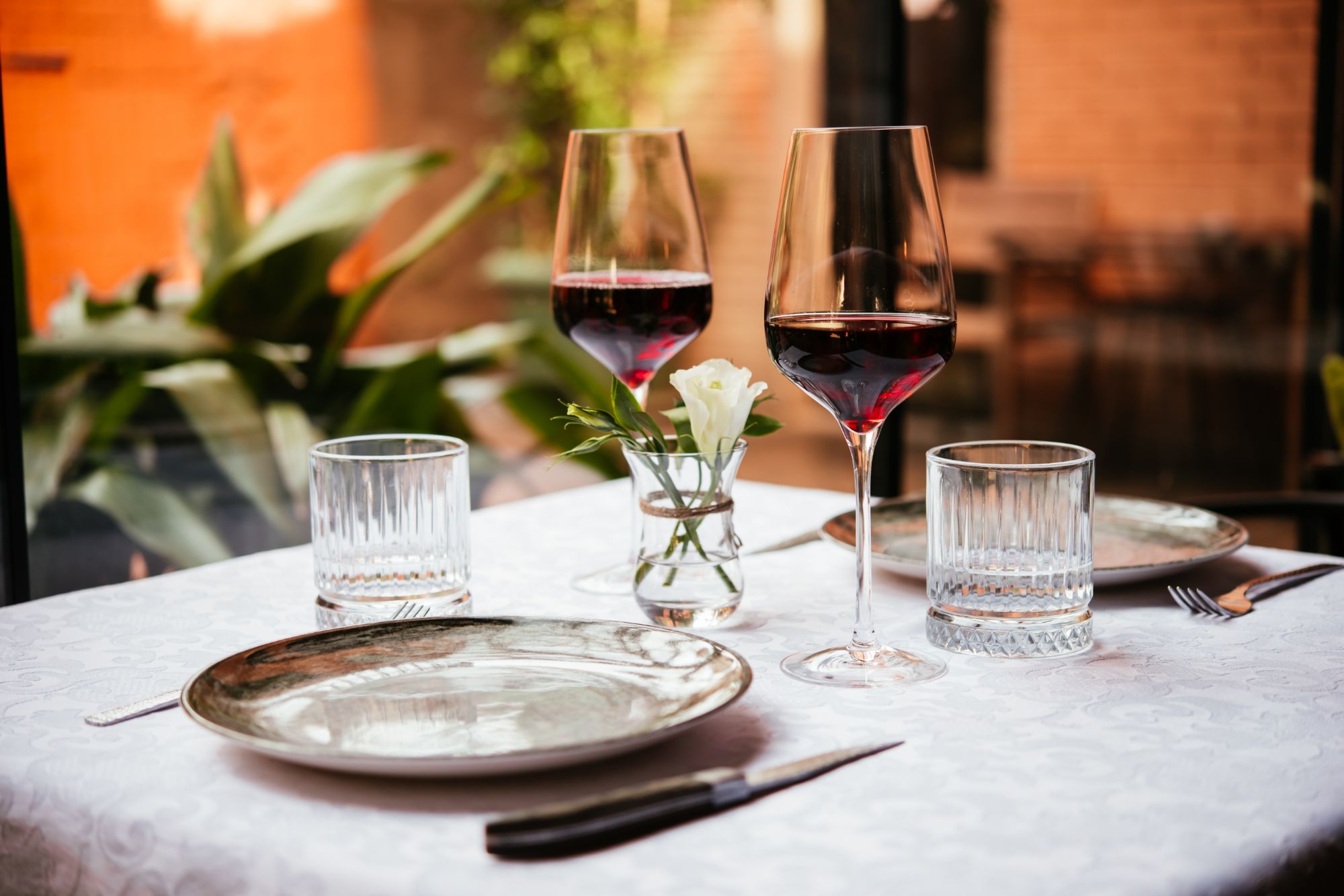 Stół w restauracji z białym obrusem, dwoma kieliszkami czerwonego wina, talerzami i kwiatem w wazonie.