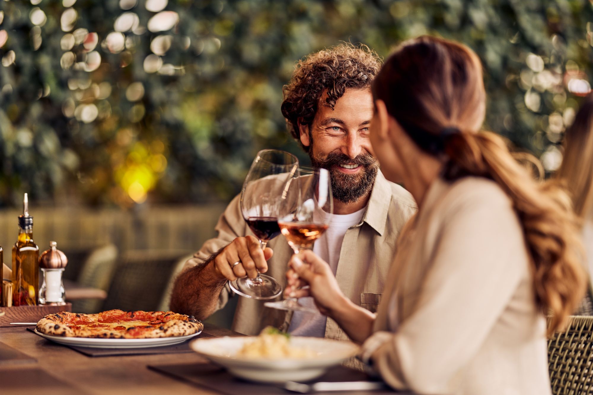 Para siedzi przy stole w restauracji, wznosząc kieliszki wina nad pizzą i makaronem.