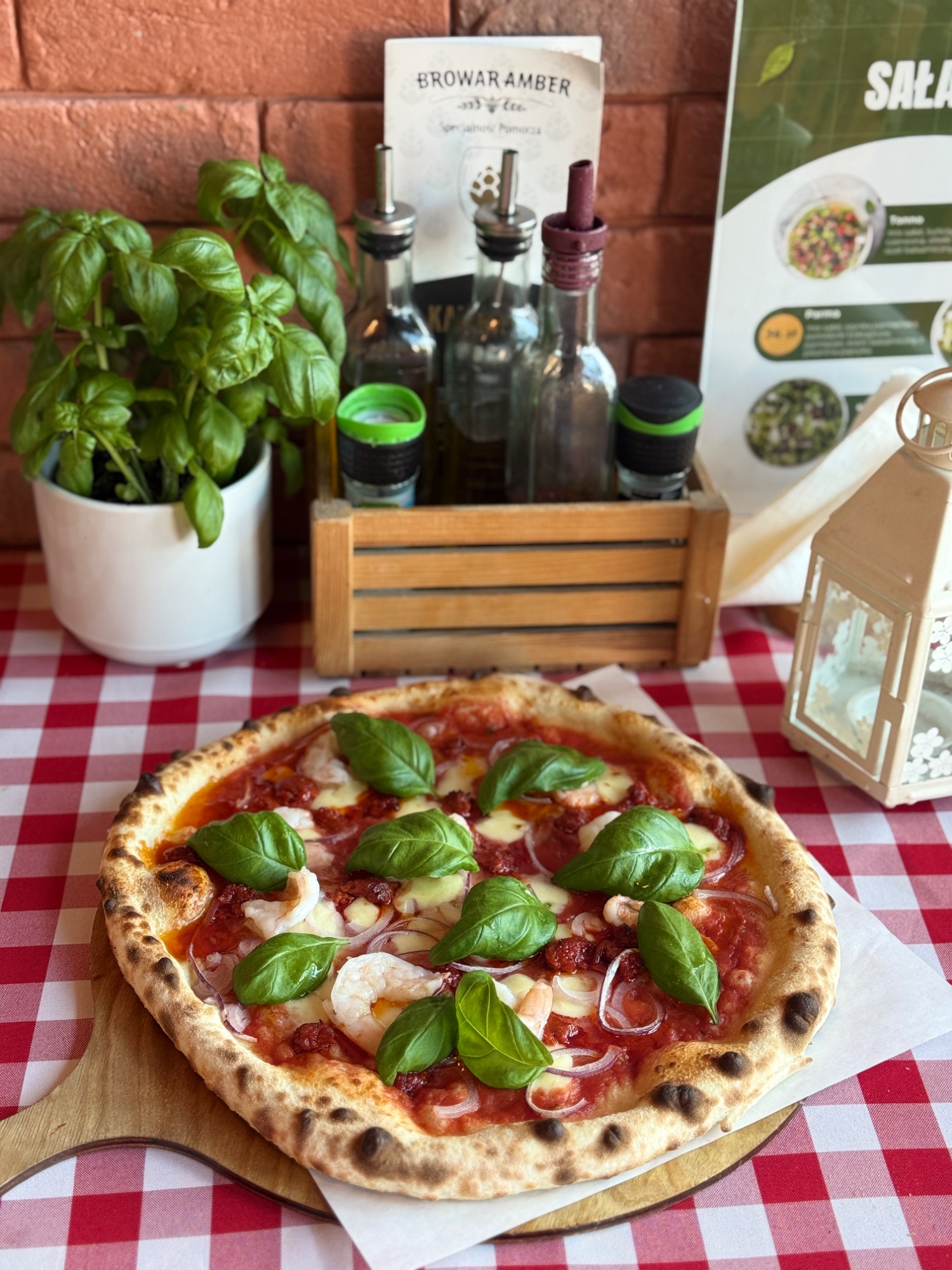 Pizza margherita z bazylią na drewnianym talerzu, zioła i butelki na tle stołu w kratkę.