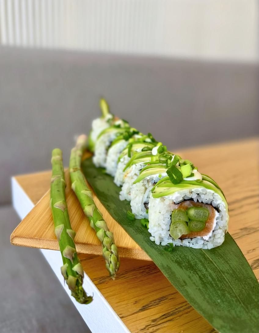 Sushi na liściu bambusa, podawane z zielonymi szparagami, udekorowane sosem i posypane szczypiorkiem.
