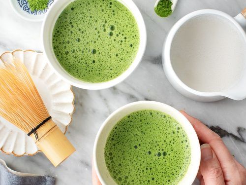 Dwie filiżanki zielonej herbaty matcha na marmurowym blacie, obok dzbanek i miotełka do matchy.