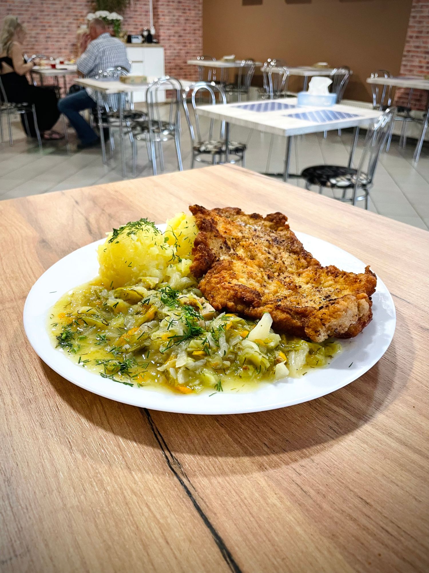 Złocista kotlet schabowy z ziemniakami i kapustą na talerzu, w restauracji z drewnianym stołem.