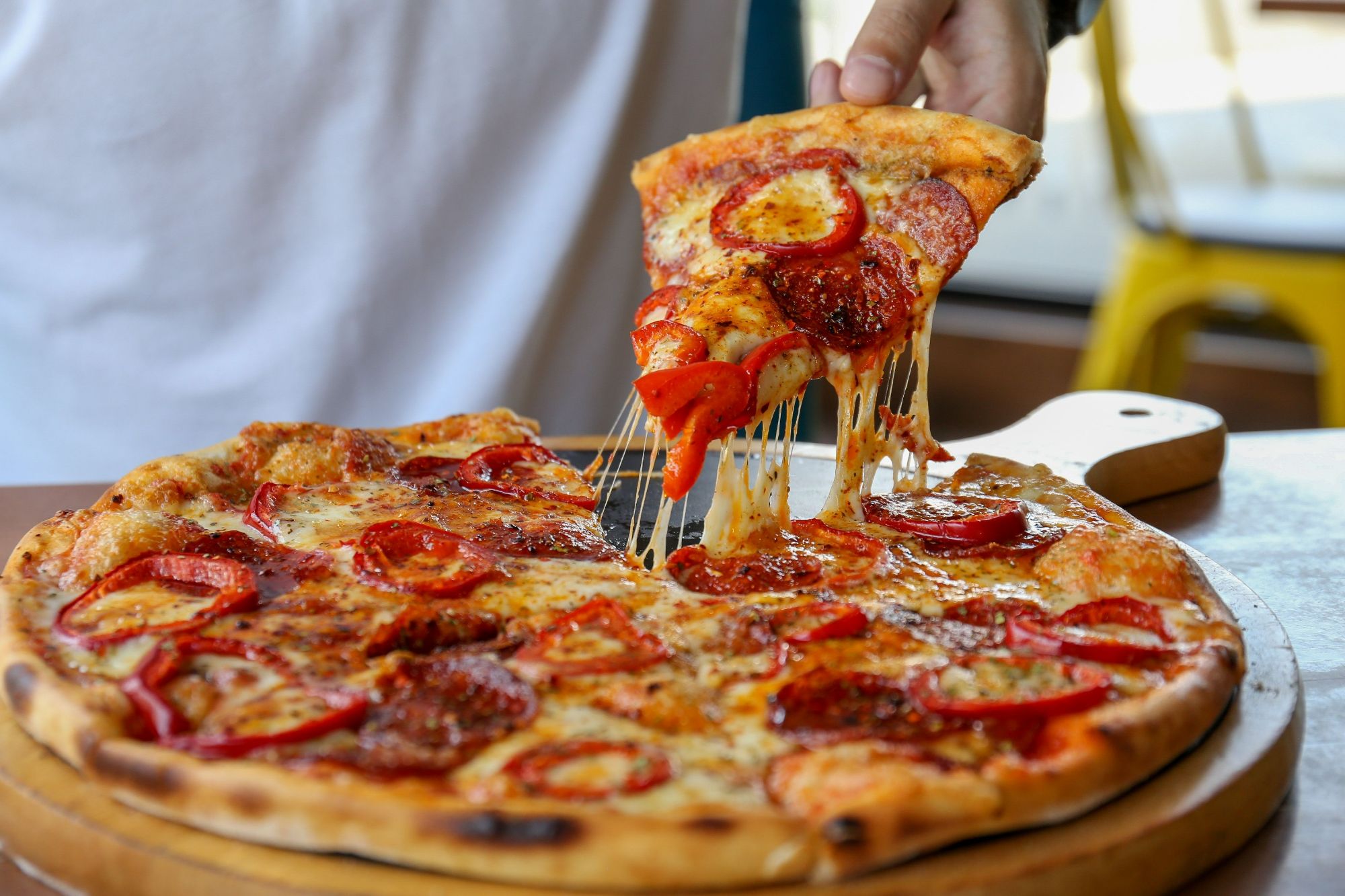 Osoba wyciąga kawałek pizzy pepperoni z serem z okrągłej deski, na tle stołu i krzesła.