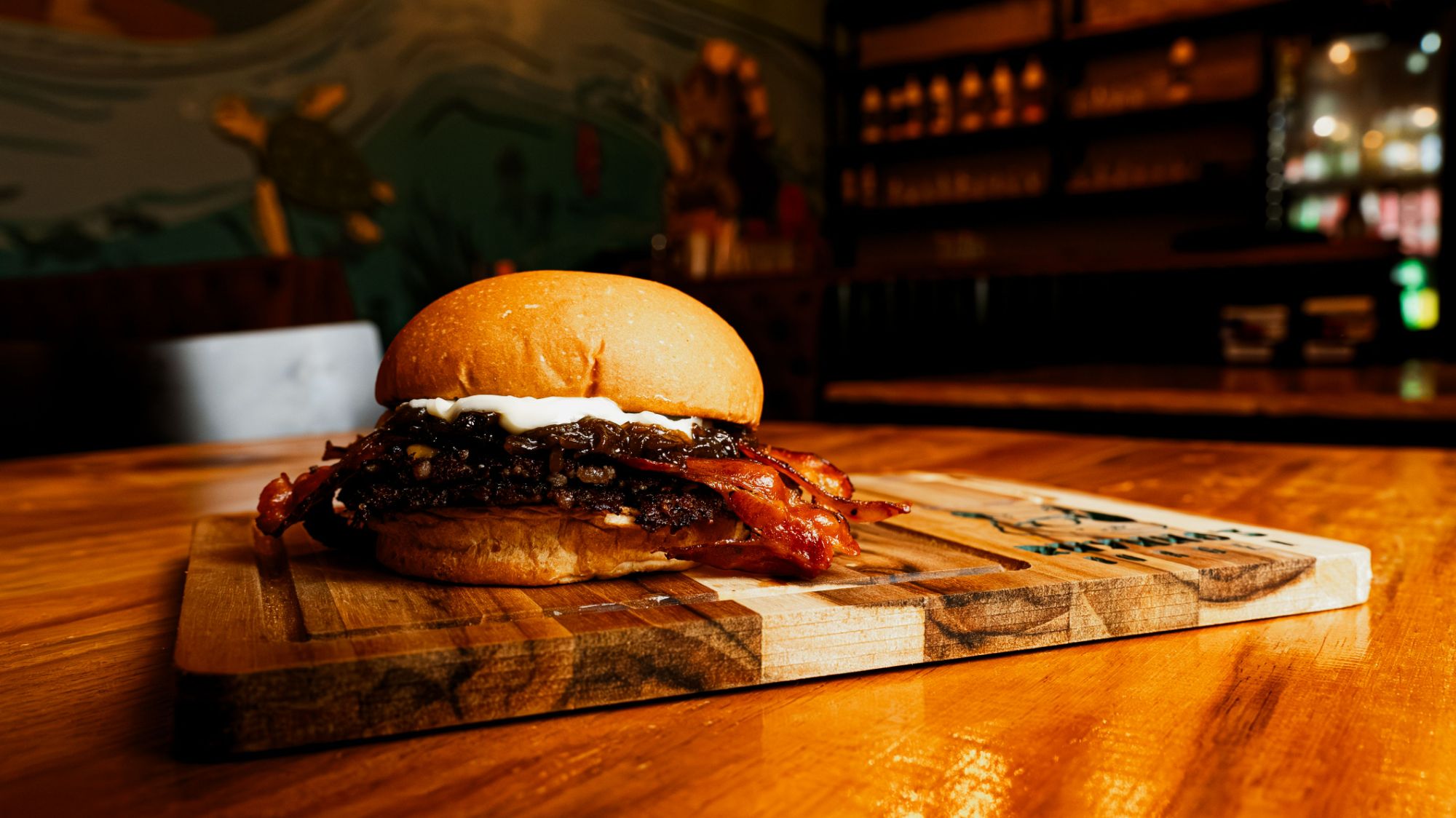 Soczysty hamburger na desce, z boczkiem i sosem, w tle przytulna restauracja.