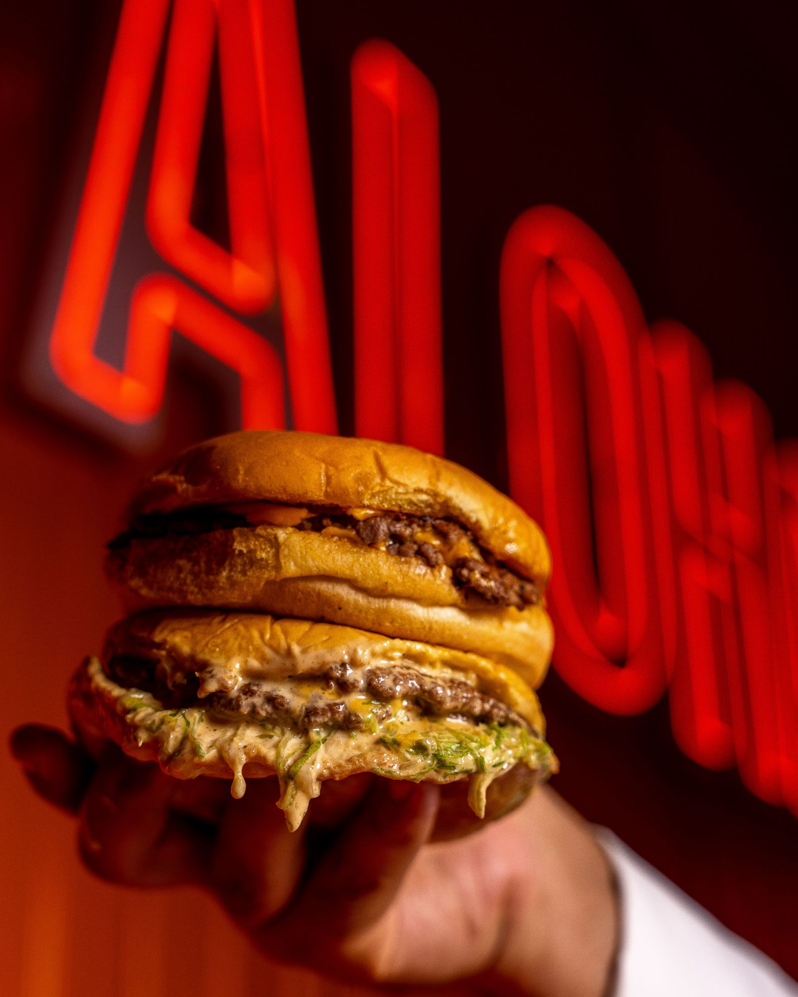 Osoba trzymająca soczystego burgera z serem i sałatą, z neonowym napisem „Aloha” w tle.