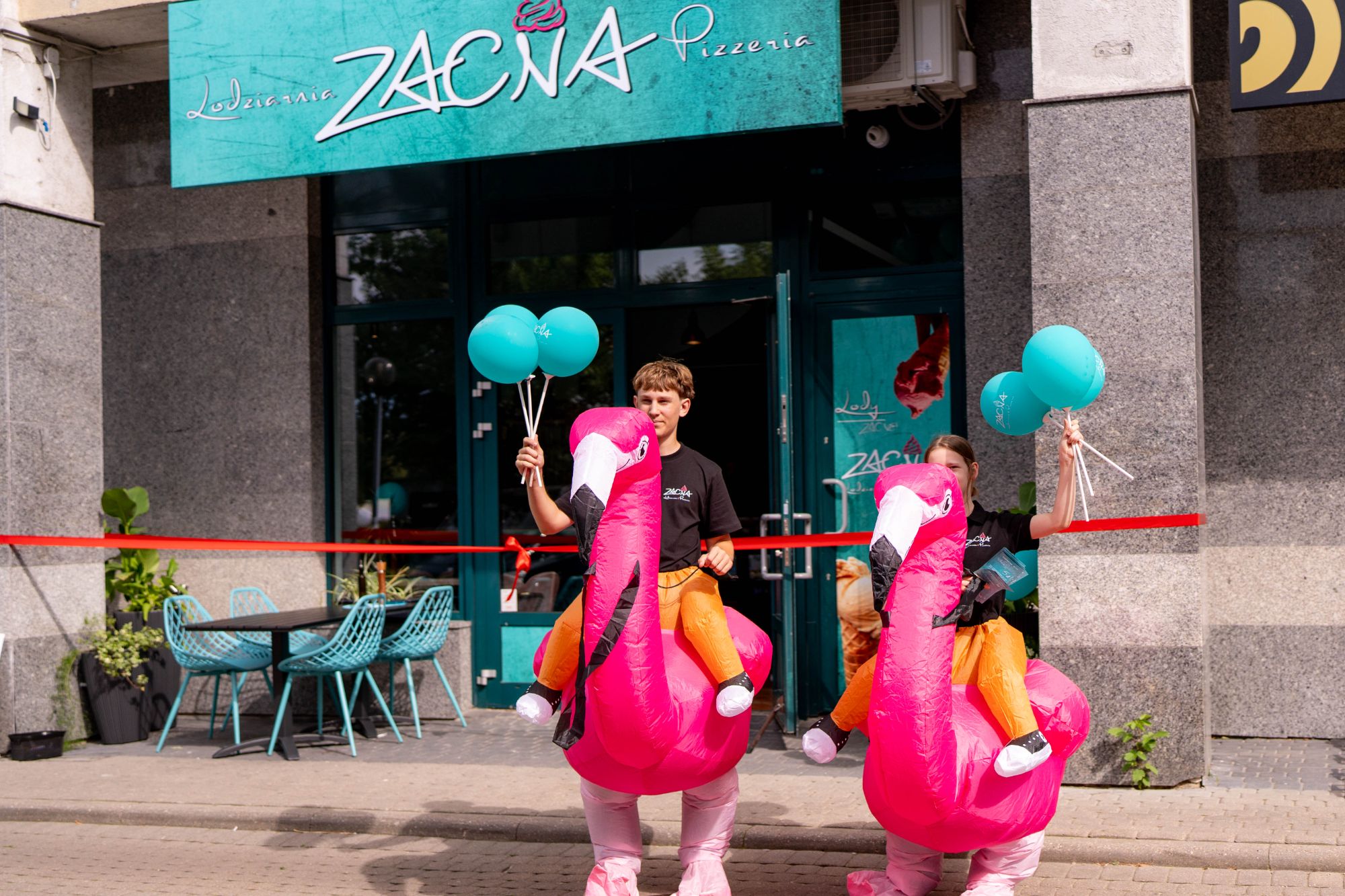 Dzieci w różowych strojach flamingów trzymają balony przed pizzerią Zacna, z czerwonymi wstęgami w tle.