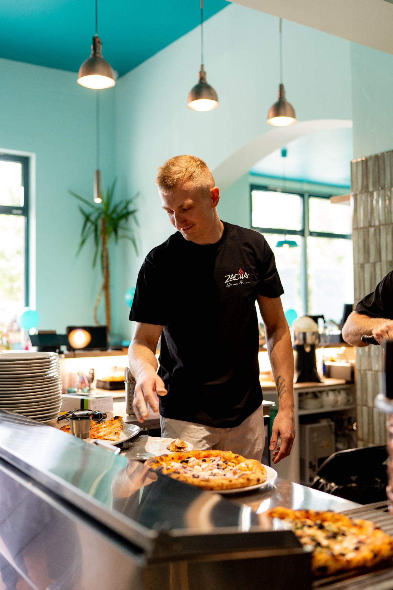 Mężczyzna w czarnej koszulce przygotowuje pizzę w nowoczesnej kuchni restauracyjnej z jasnym wystrojem.