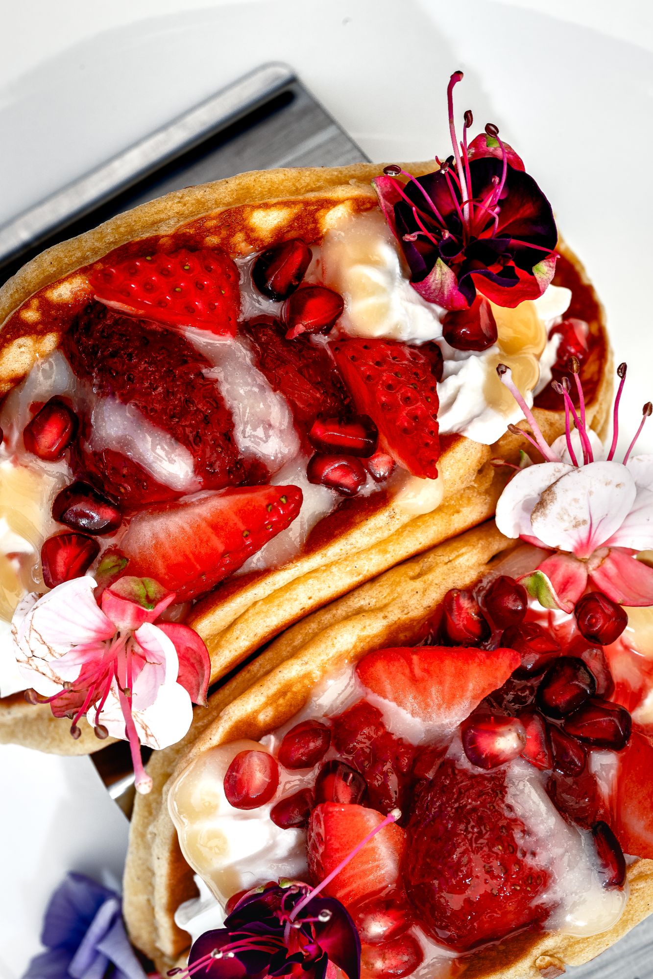 Pancakes z kremem, truskawkami, granatem i jadalnymi kwiatami na białym talerzu.