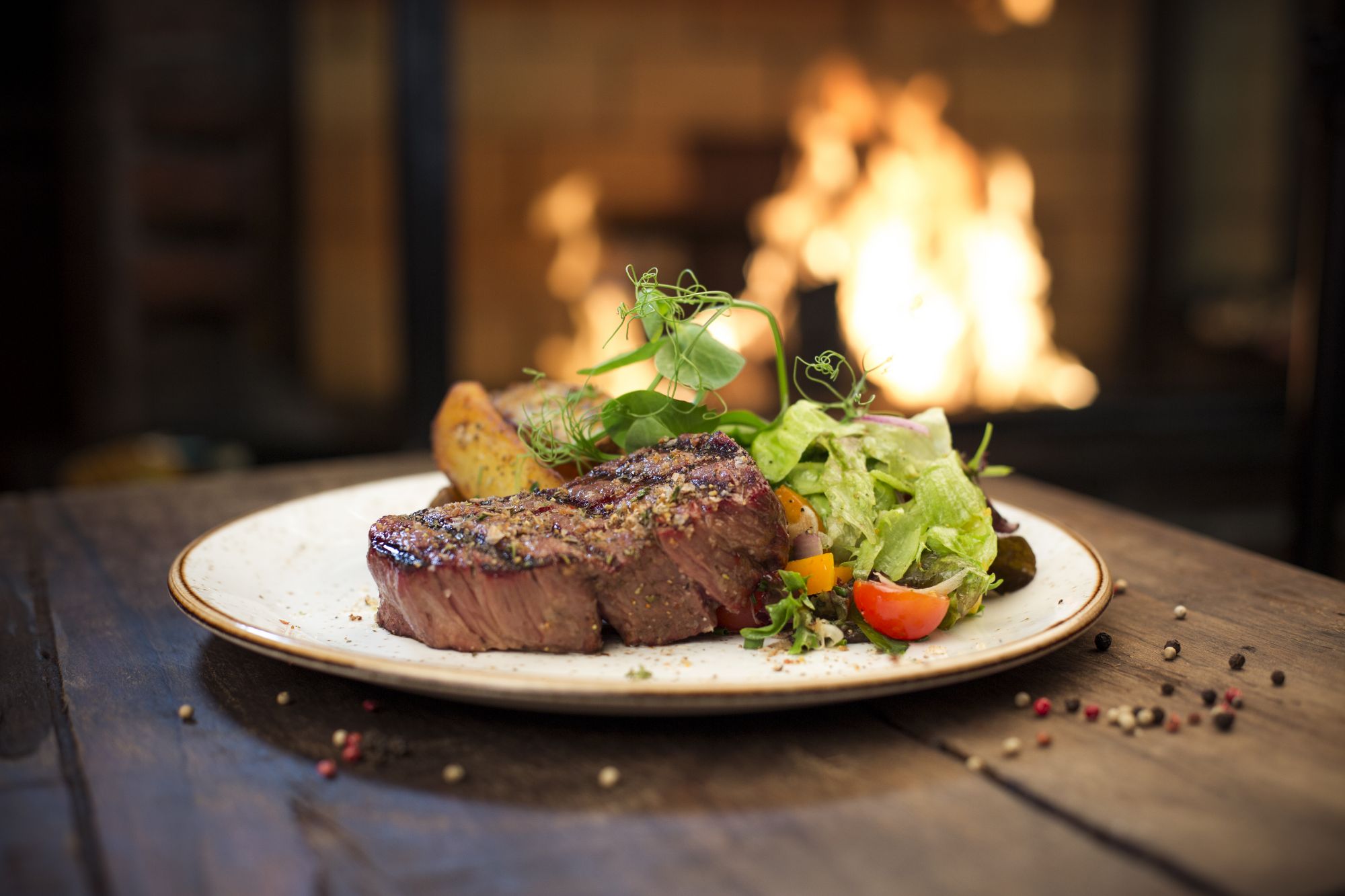 Steak z sałatką i ziemniakami na talerzu, w tle palący się kominek.
