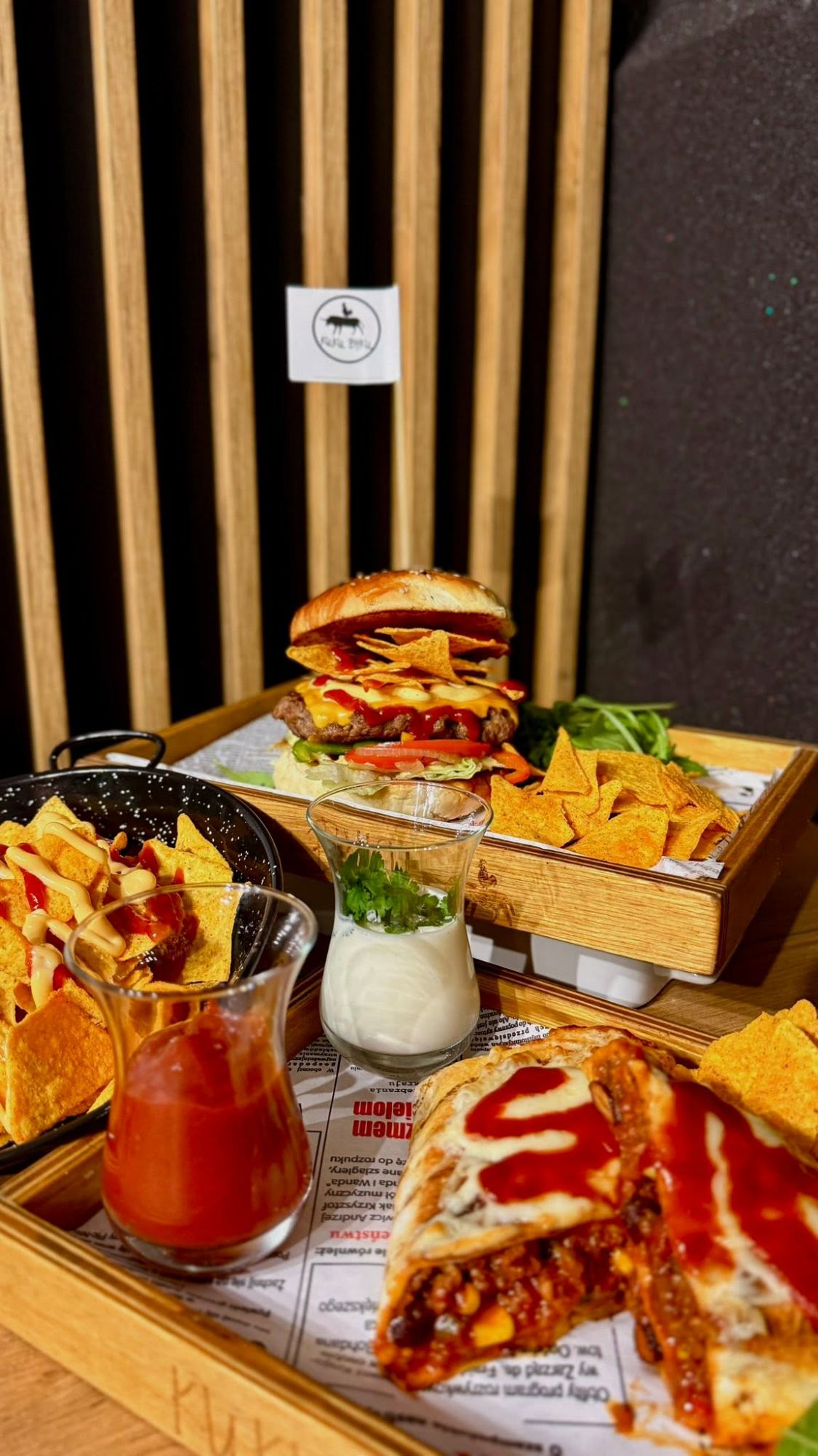 Taca z jedzeniem: burger z frytkami, sos chili, nachos i deser w szklanej miseczce.