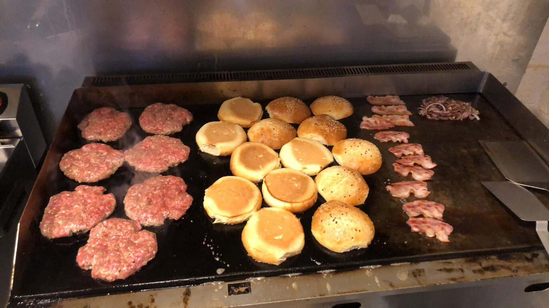 Grill z hamburgerami, bułkami i boczkiem, parujące świeże składniki na rozgrzanej płycie.