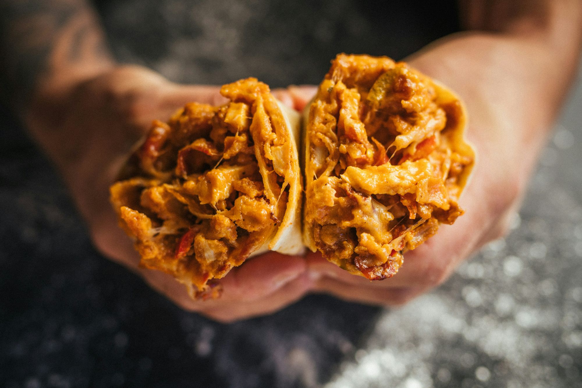 Dwie połówki burrito z mięsem i warzywami trzymane w dłoniach na tle ciemnego blatu.