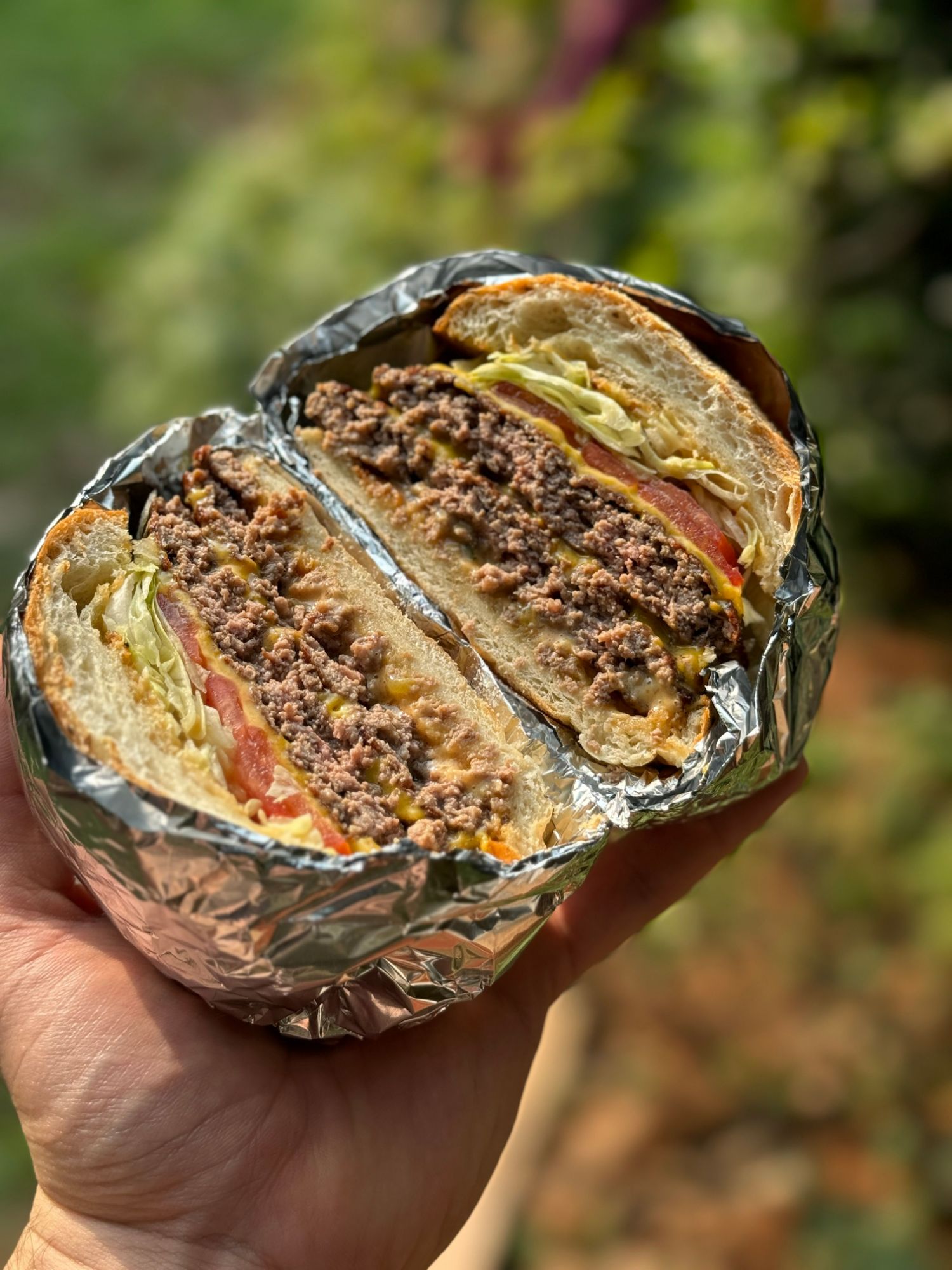 Burger z mielonym mięsem, sałatą, pomidorem, zawinięty w folię, trzymany w ręku na tle zieleni.