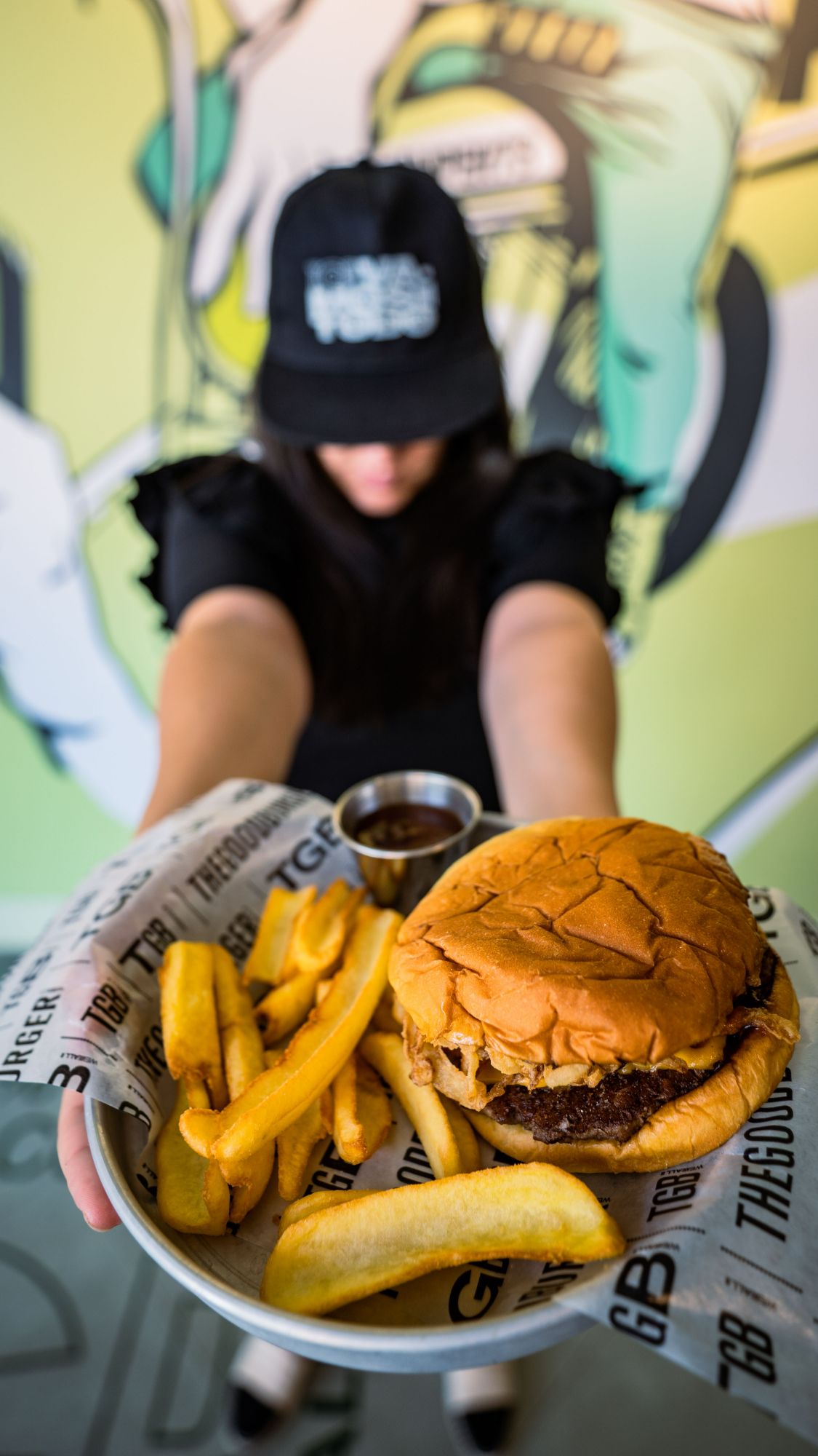 Kobieta trzyma talerz z burgerem i frytkami przed kolorową ścianą w lokalu gastronomicznym.