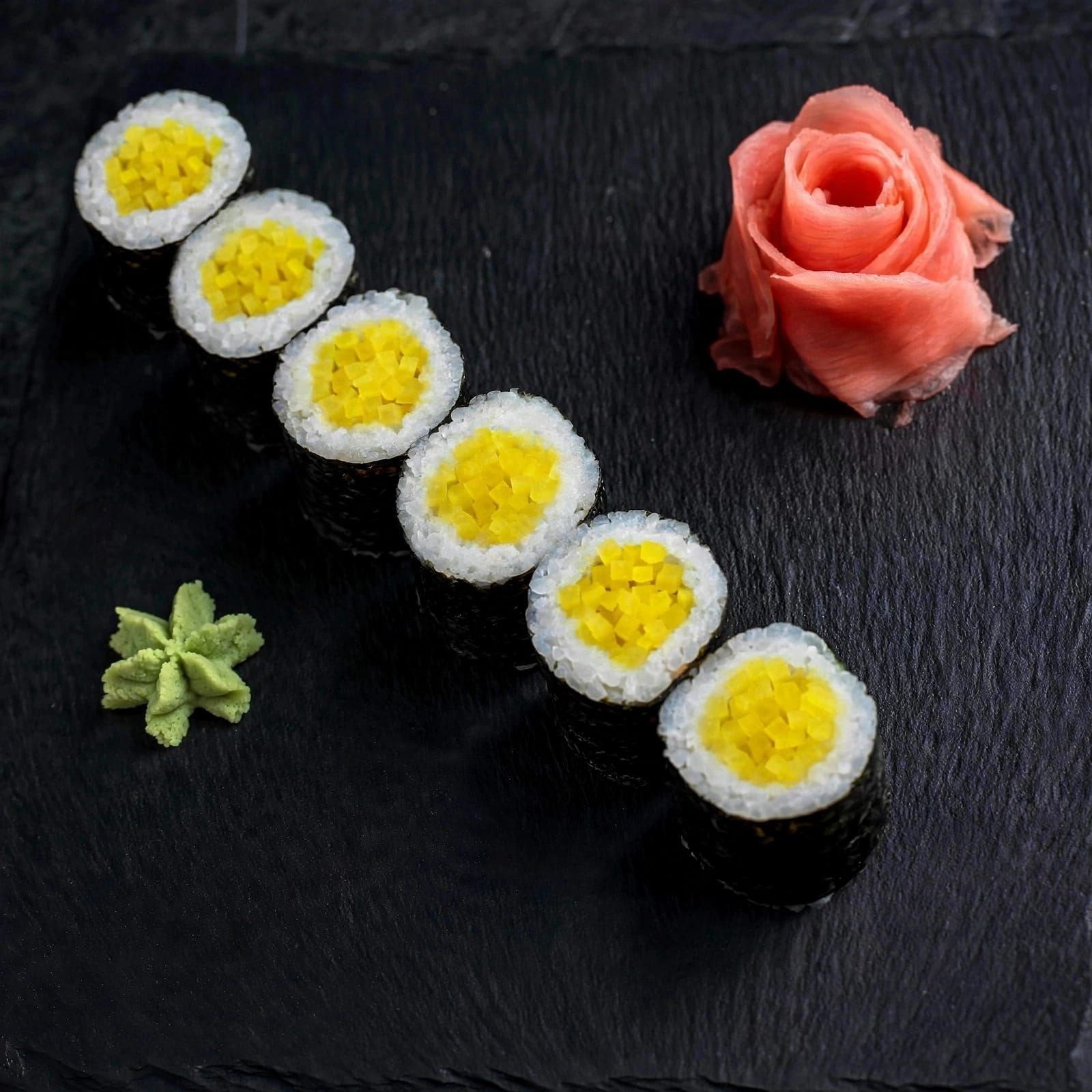 Sushi z żółtym kwadratem nadzienia, podane na czarnej płycie, z różą z różowego imbiru i wasabi obok.