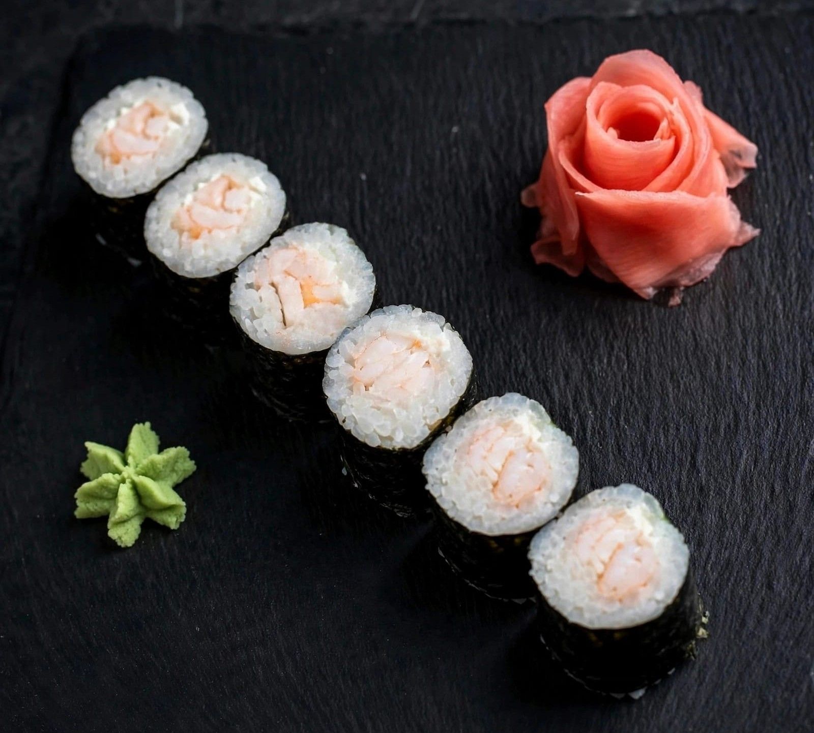 Sushi rolki z ryżem i łososiem, podane z różą imbirową i wasabi na czarnej płycie.