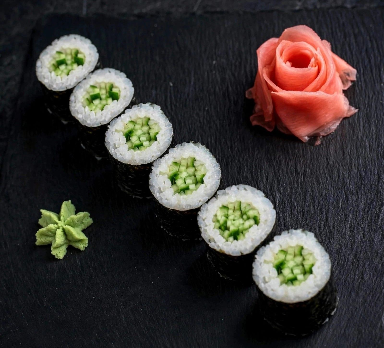Sześć rolek sushi z ogórkiem, podane z różową różą z imbiru i wasabi na czarnym talerzu.