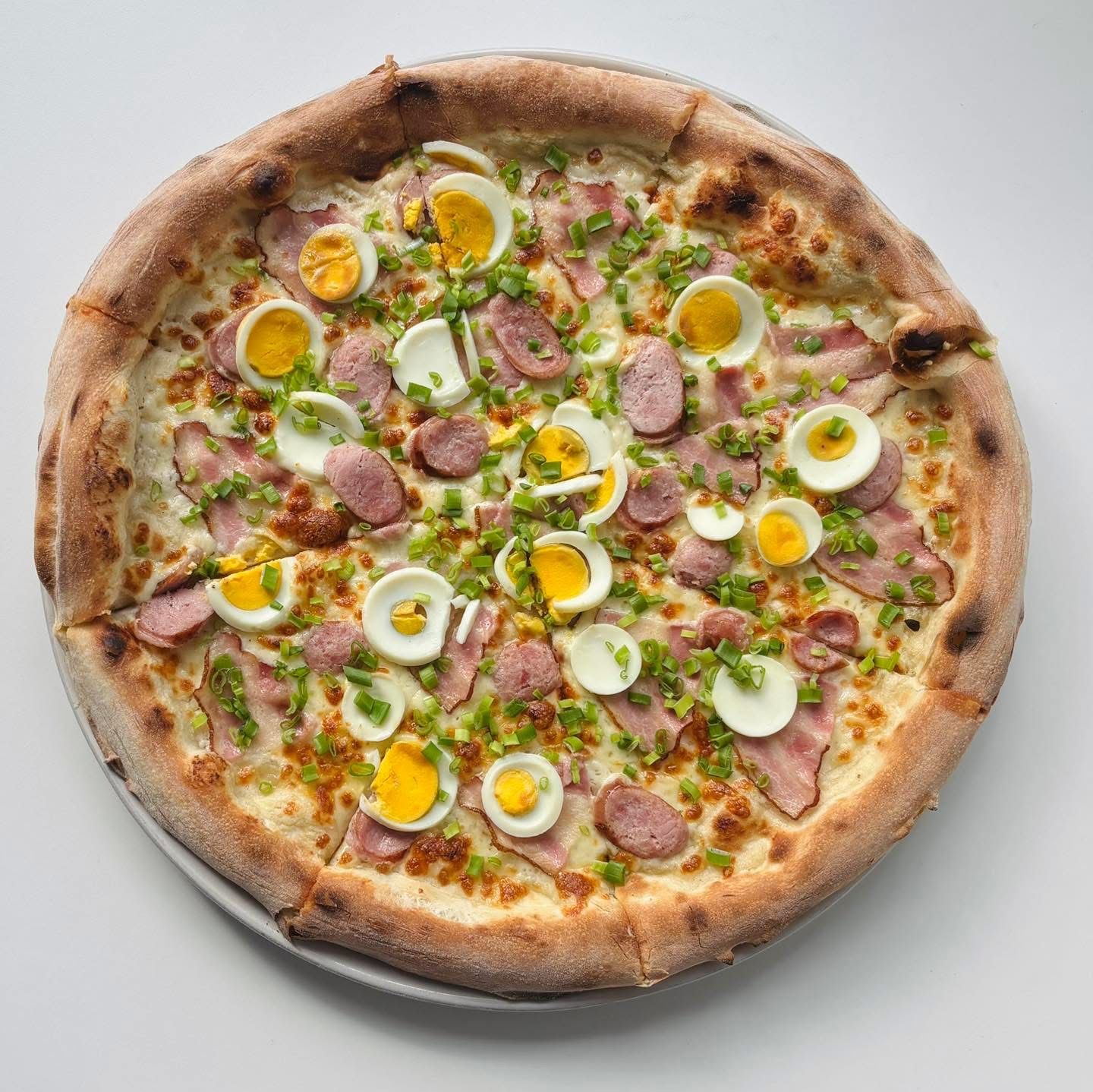 Pizza z jajkiem, kiełbasą, szczypiorkiem i serem na cienkim cieście, podana na talerzu.