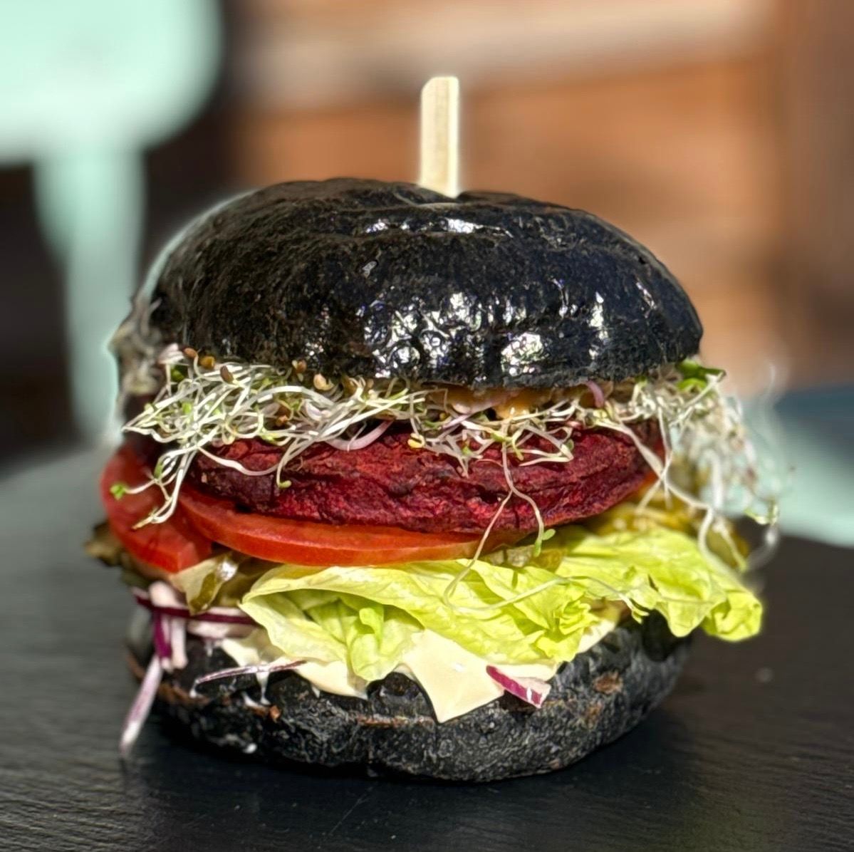 Burger z czarną bułką, buraczanym kotletem, sałatą, pomidorem i kiełkami na tle nieostrego tła.