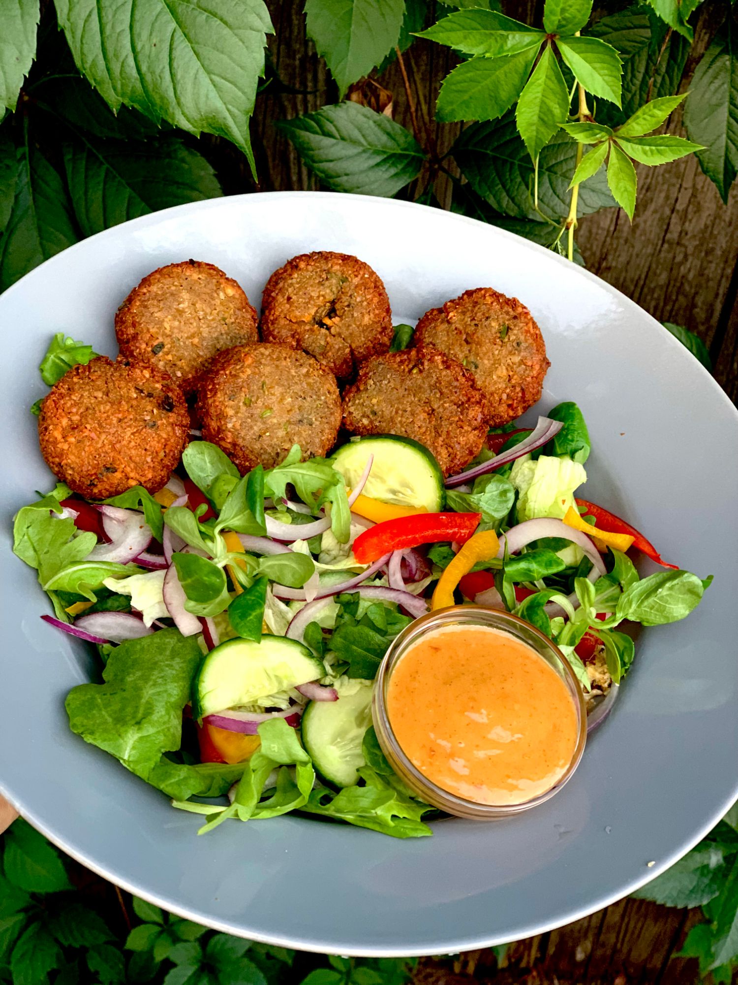Sałatka z kolorowymi warzywami, chlebki falafel i sos w szklanym naczyniu, podana na tle zielonych liści.