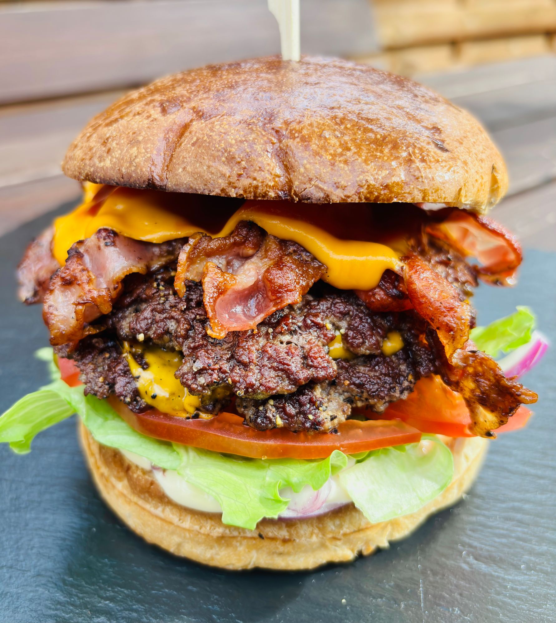 Burger z wołowiną, bekonem, serem cheddar, sałatą i pomidorem na bułce pełnoziarnistej, podany na czarnym talerzu.