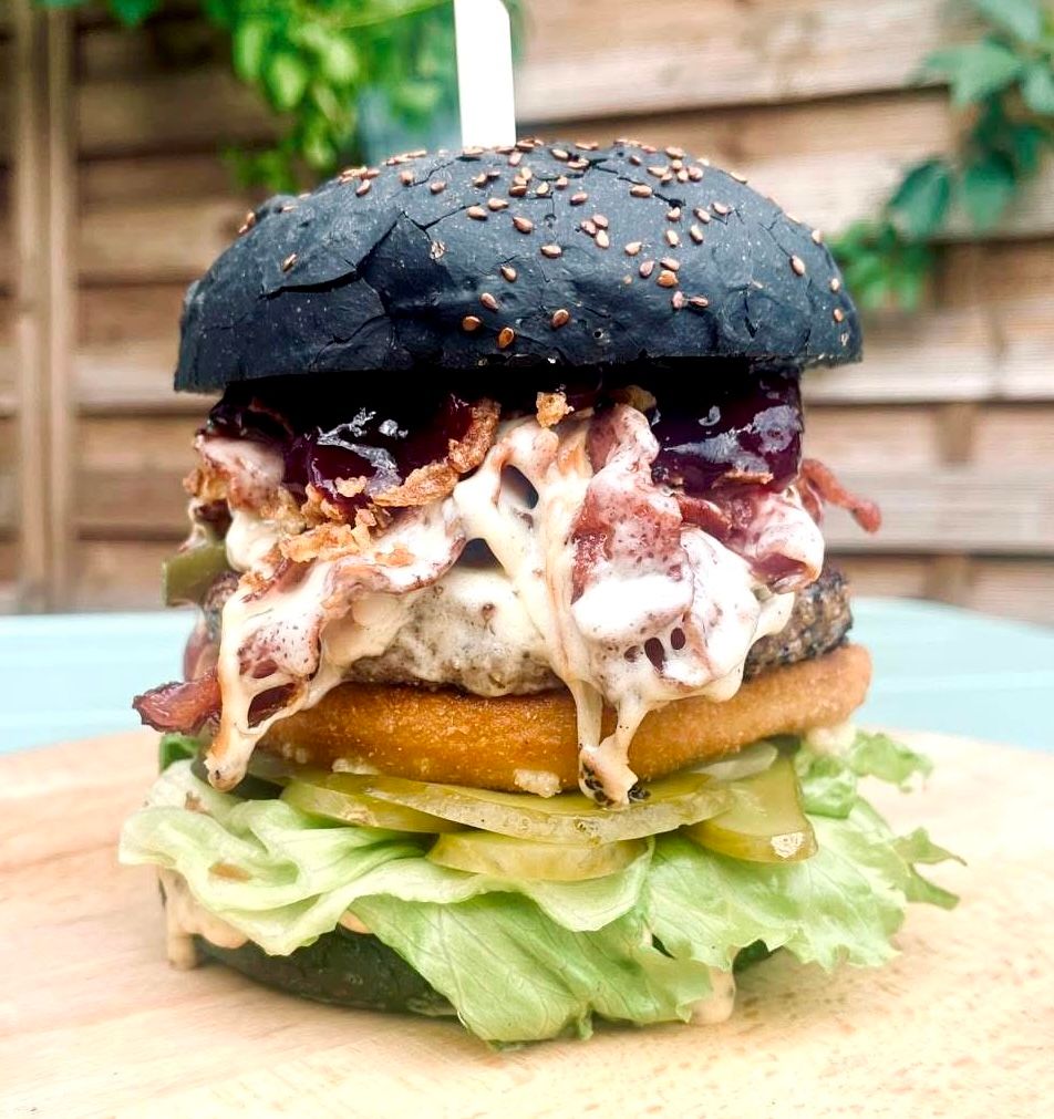 Czarny burger z wołowiną, boczkiem, sałatą, piklami, sosem i chrupiącą bułką na drewnianym talerzu.