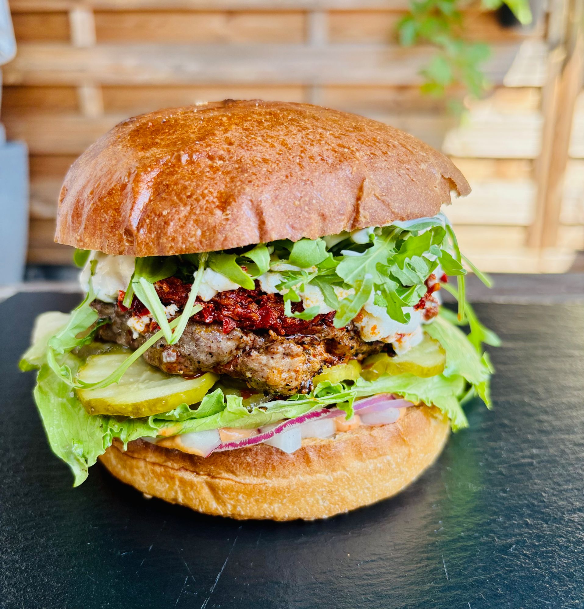 Soczysty hamburger w bułce, z sałatą, ogórkiem, cebulą i sosami, podany na ciemnym tle.