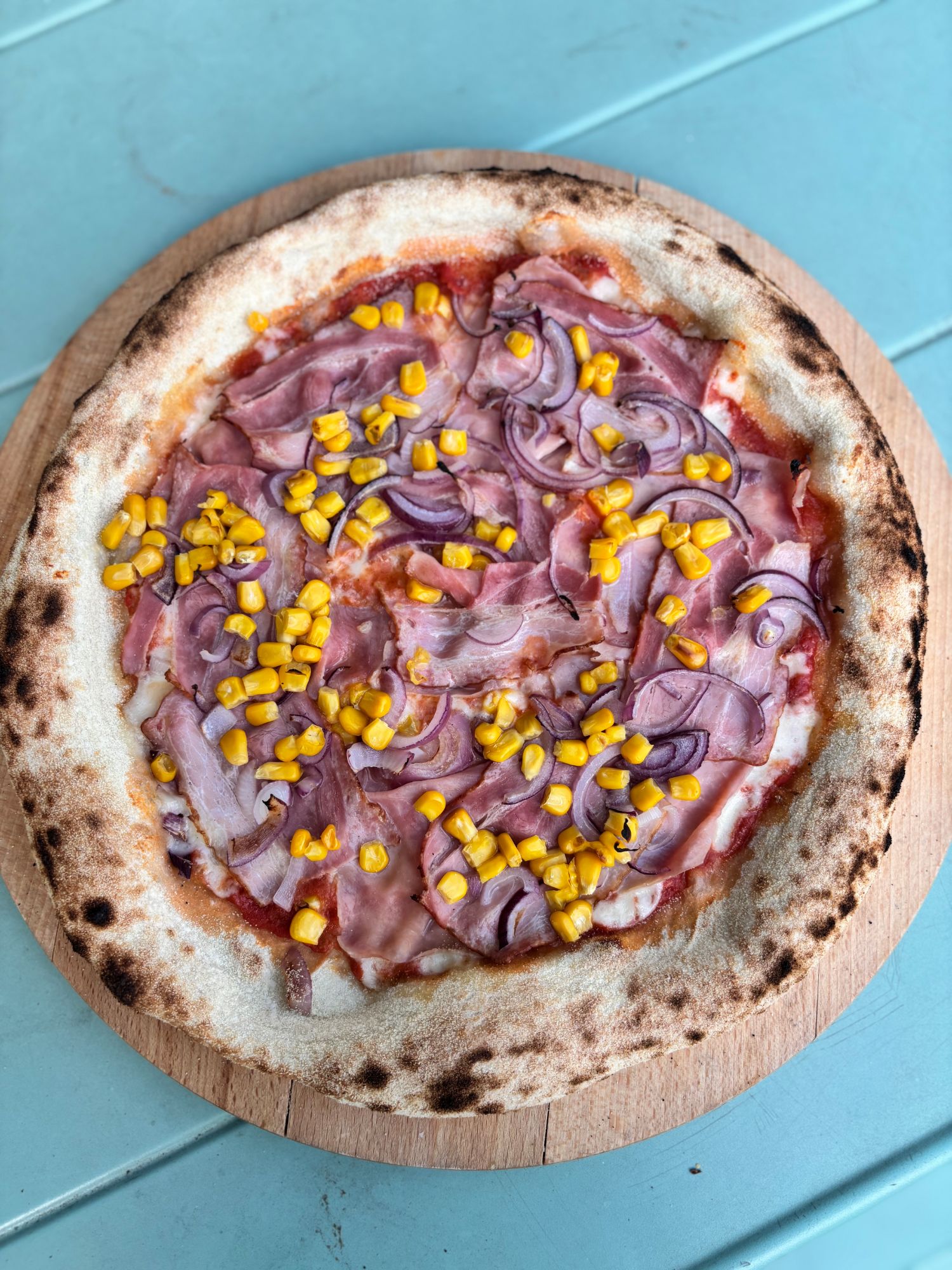 Pizza z szynką, kukurydzą i cebulą na drewnianej desce.