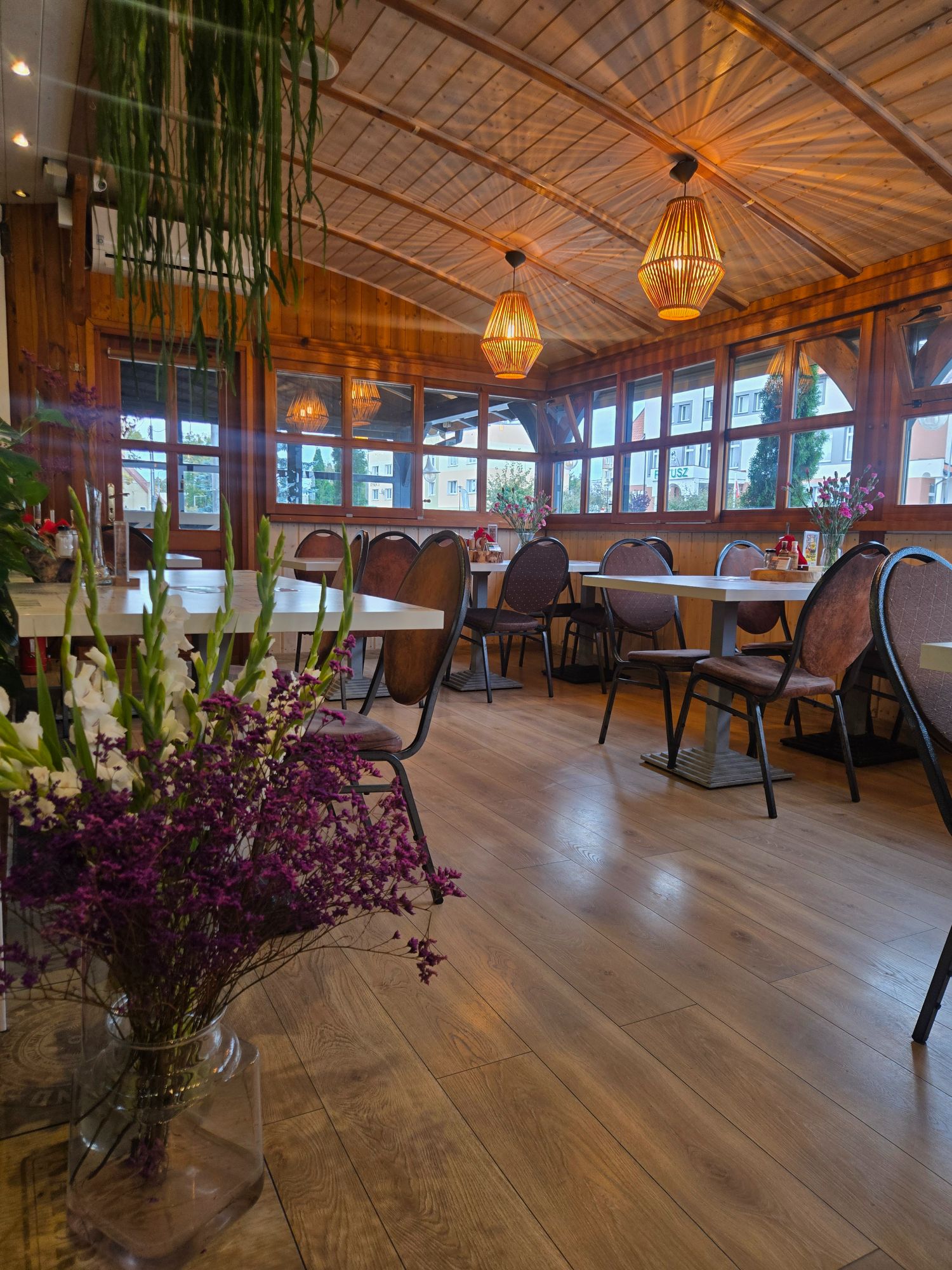 Nowoczesna restauracja z drewnianym wystrojem, dużymi oknami i stołami, ozdobiona świeżymi kwiatami.