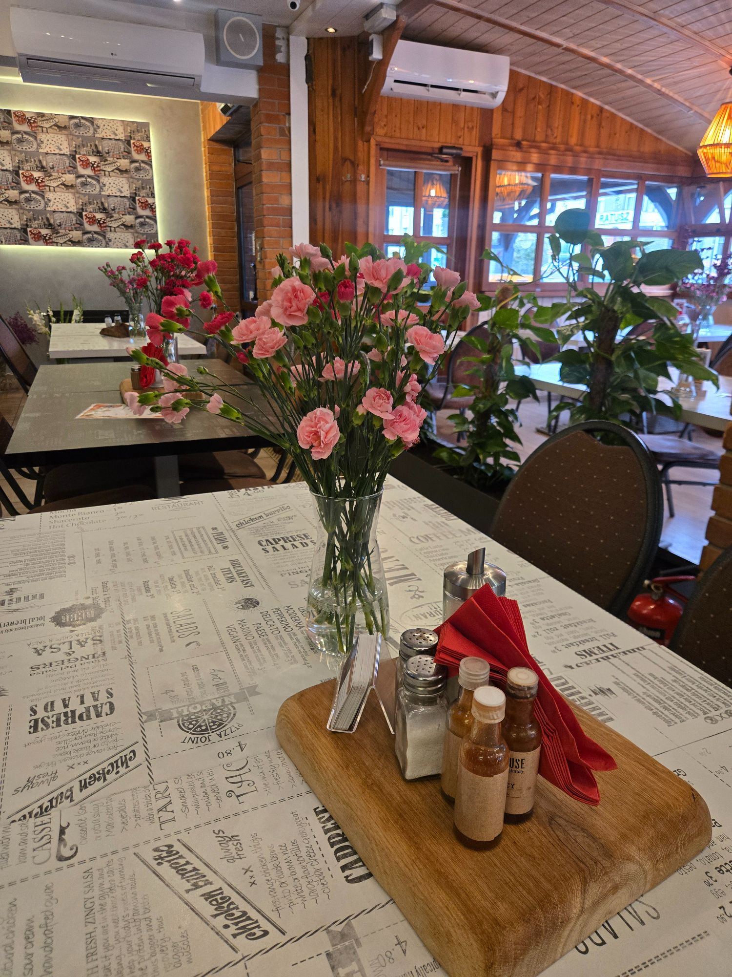 Bukiet różowych goździków w wazonie na stole w restauracji, obok przyprawy i serwetki w drewnianym zestawie.