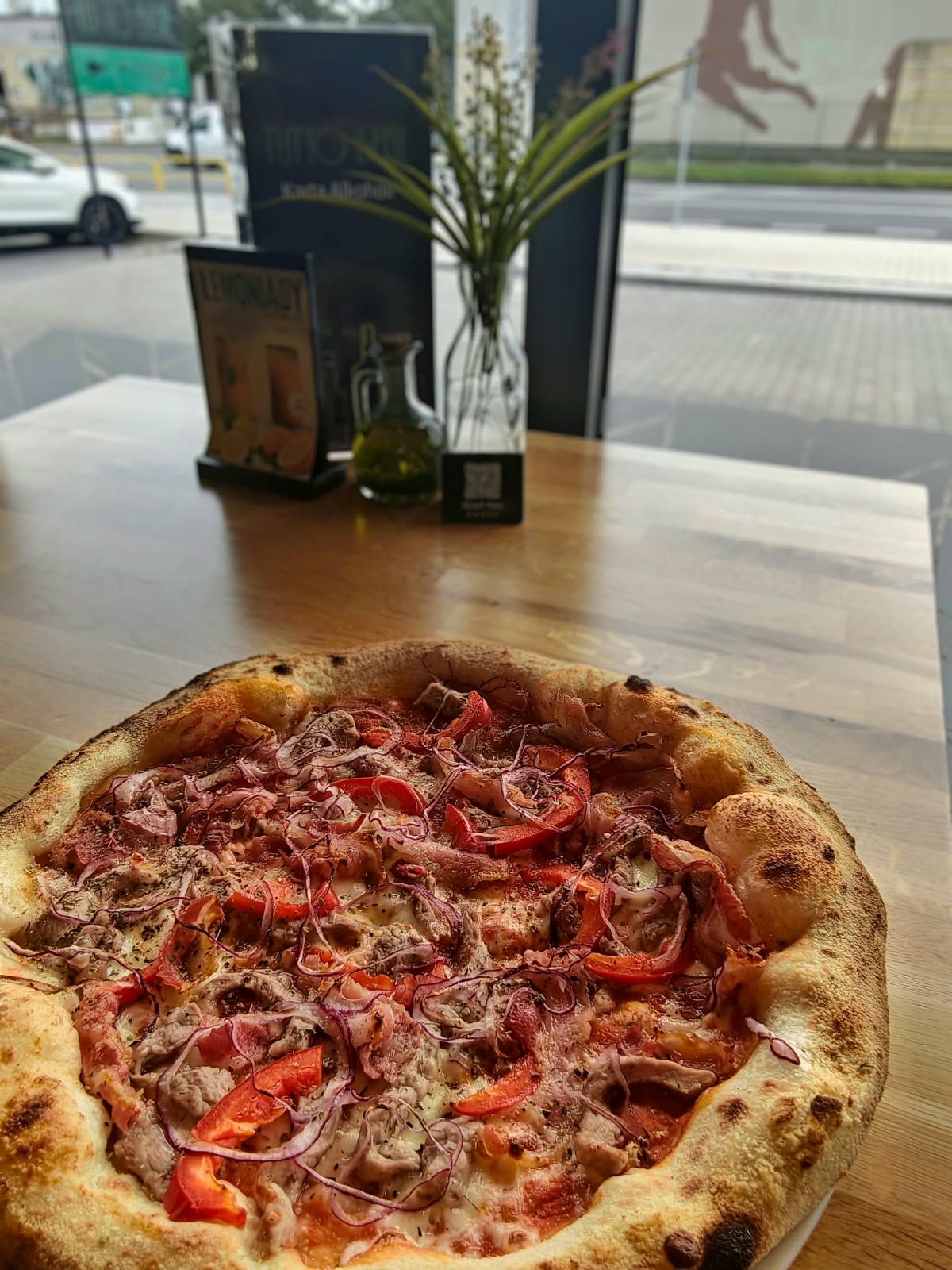 Pizza na drewnianym stole przed oknem, z dodatkami takimi jak mięso, czerwona cebula i pomidory.