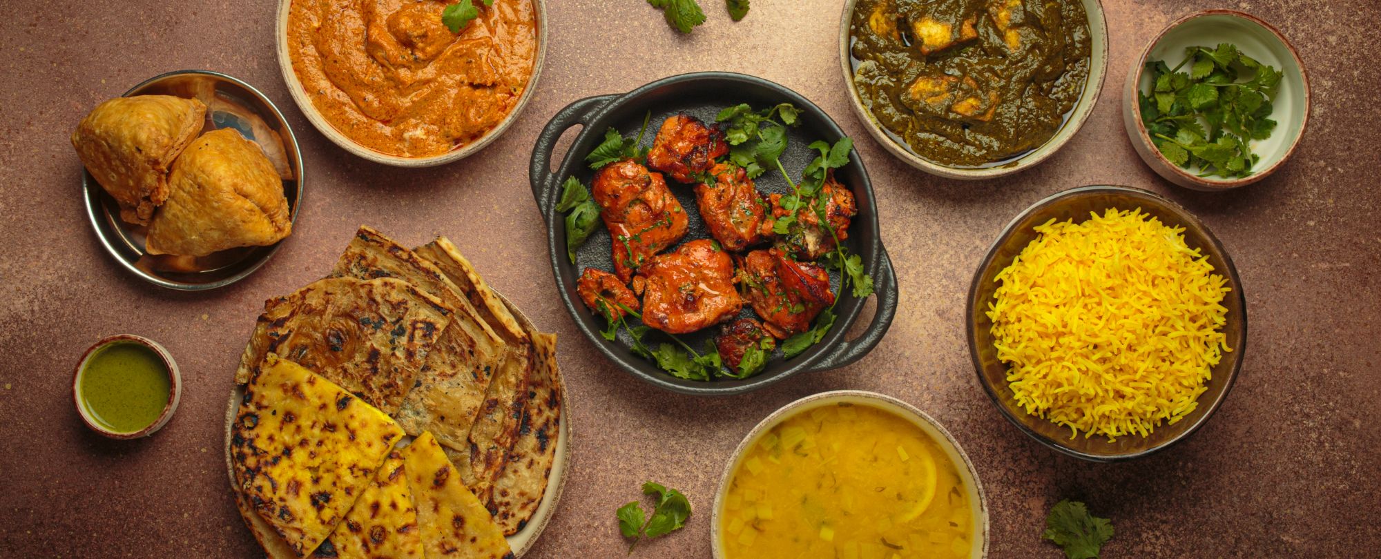 Różnorodne potrawy indyjskie: curry, ryż, chlebek naan, samosy, sałatka i przyprawy na brązowym tle.