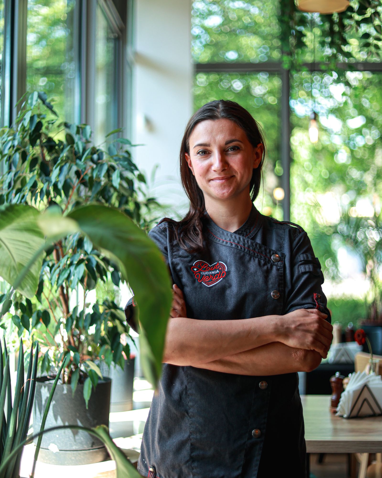 Kobieta w kucharskim fartuchu stoi z założonymi rękami w jasnej restauracji otoczonej roślinami.