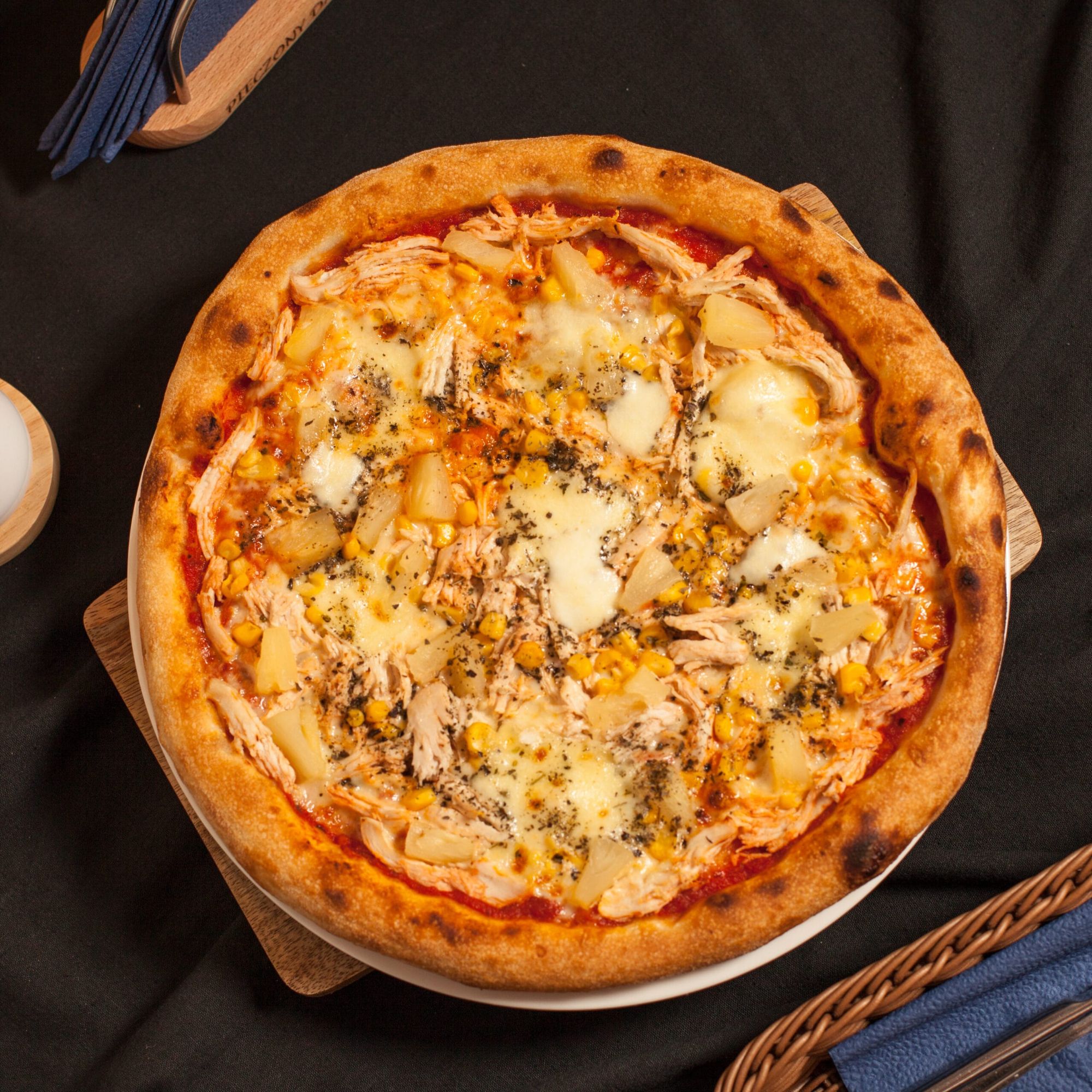Pizza z kurczakiem, ananasem i serem, na czarnym tle, podana na drewnianej desce.
