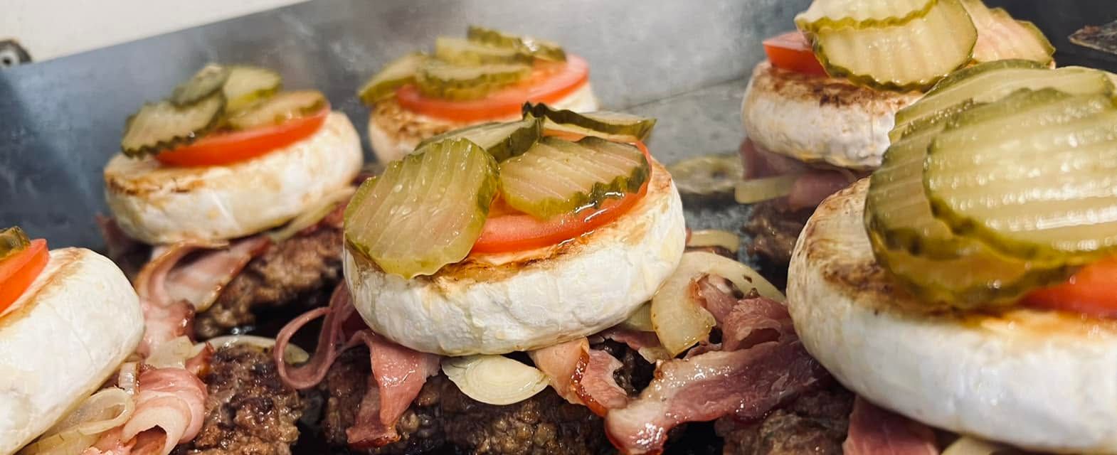 Smażone burgery z serem, cebulą, pomidorem i ogórkiem na patelni.