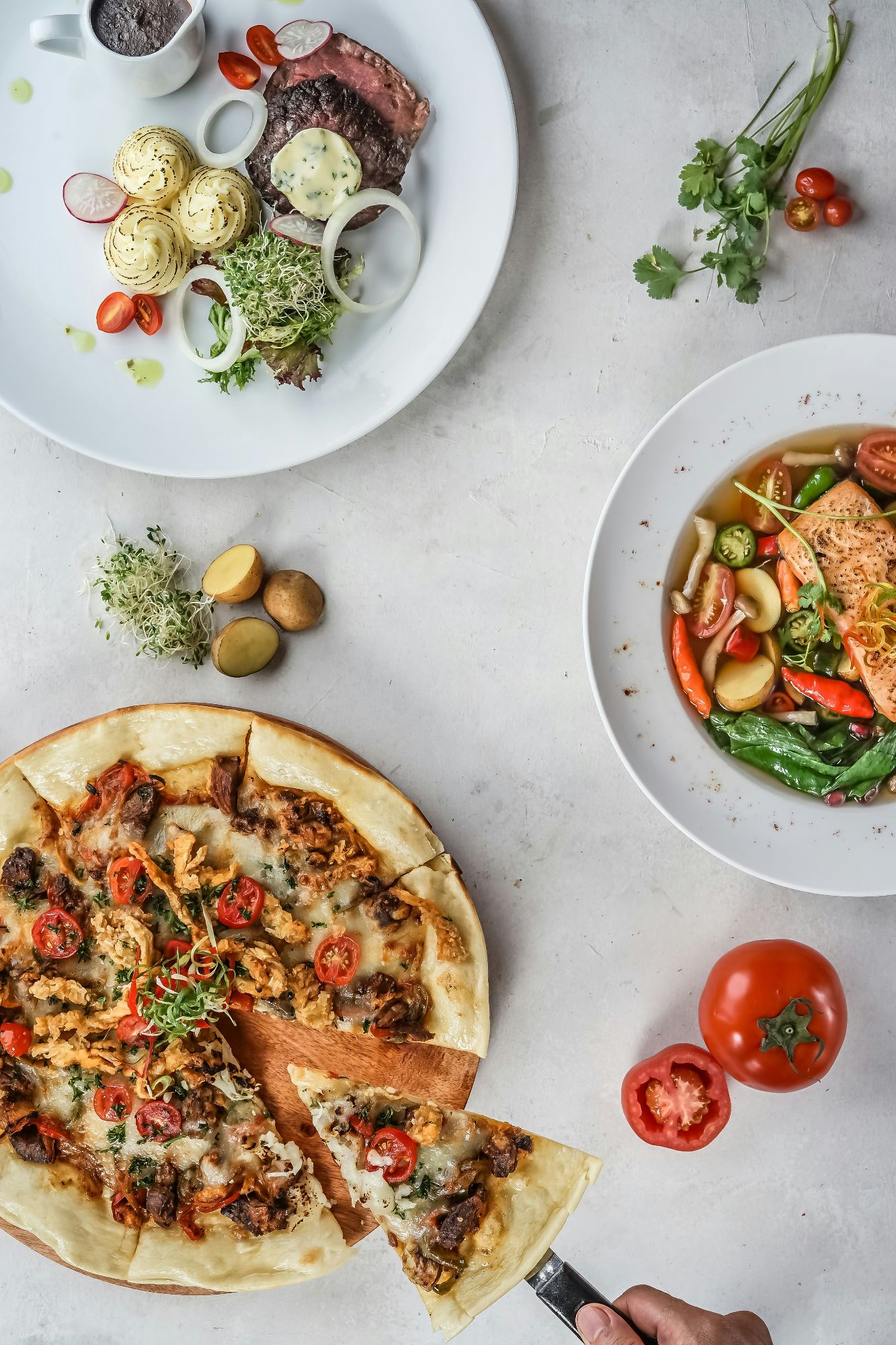 Diverse preparate culinare pe masă: pizza cu roșii, supă cu somon și fel de mâncare cu carne și garnituri.