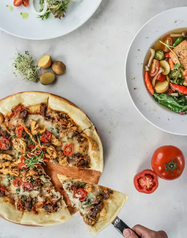 Diverse preparate culinare pe masă: pizza cu roșii, supă cu somon și fel de mâncare cu carne și garnituri.